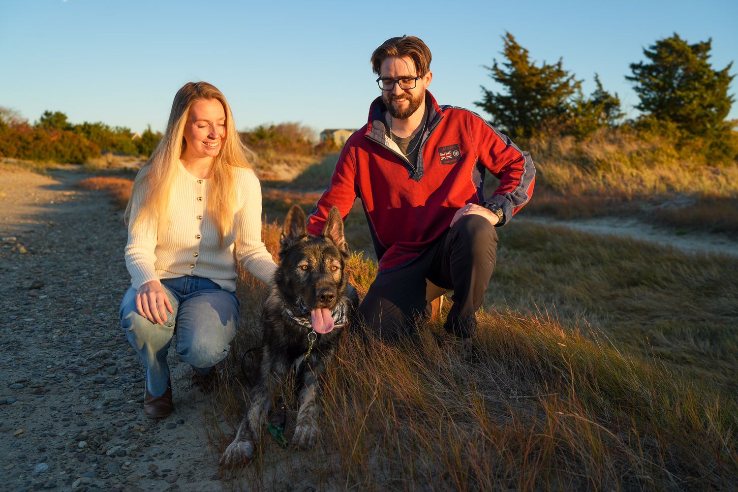 Couple kneeling beside their German Shepherd dog on a sunny beach trail with golden grass.