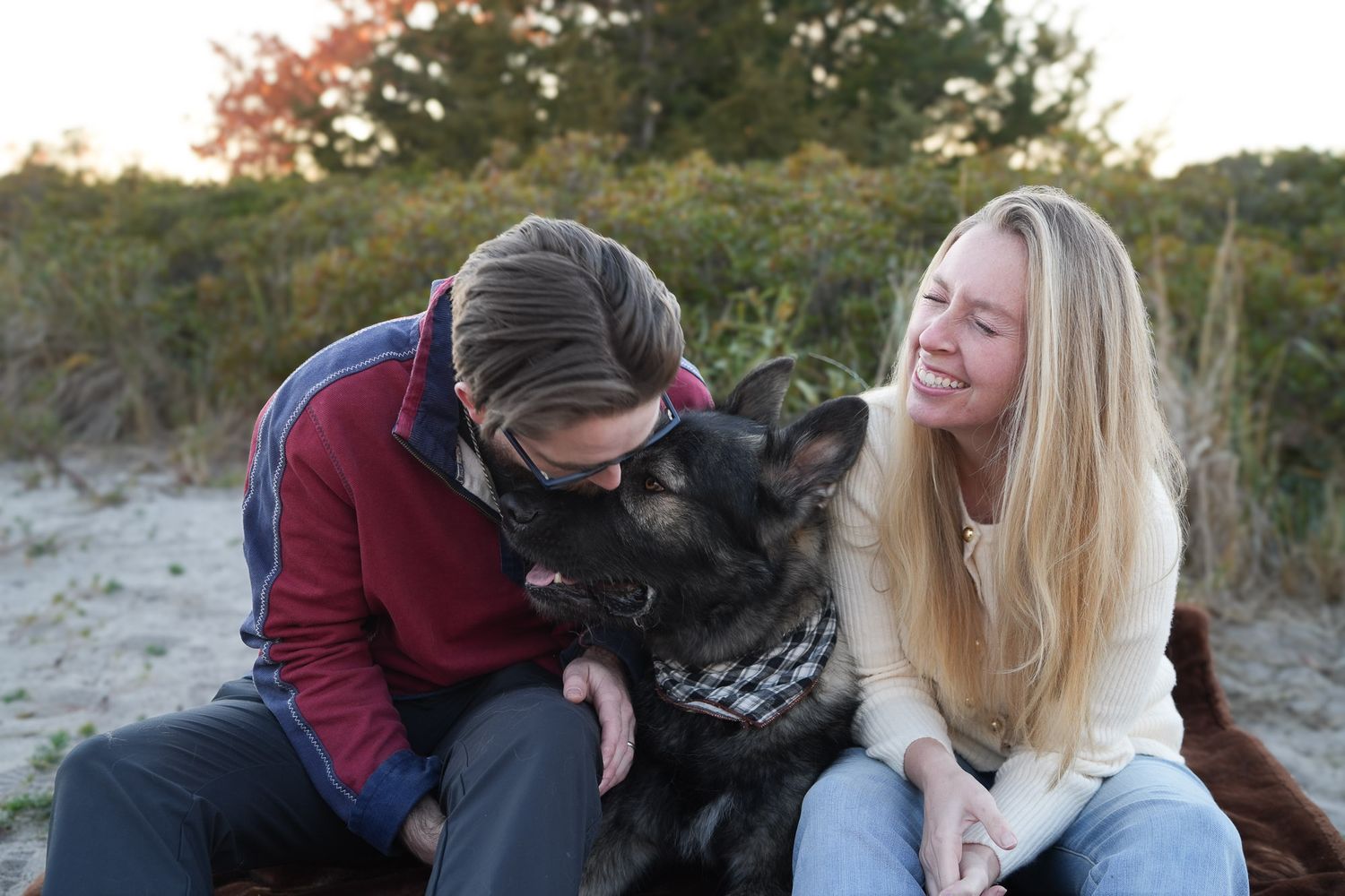 Laughing couple cuddling with their happy German Shepherd dog on a sandy beach.