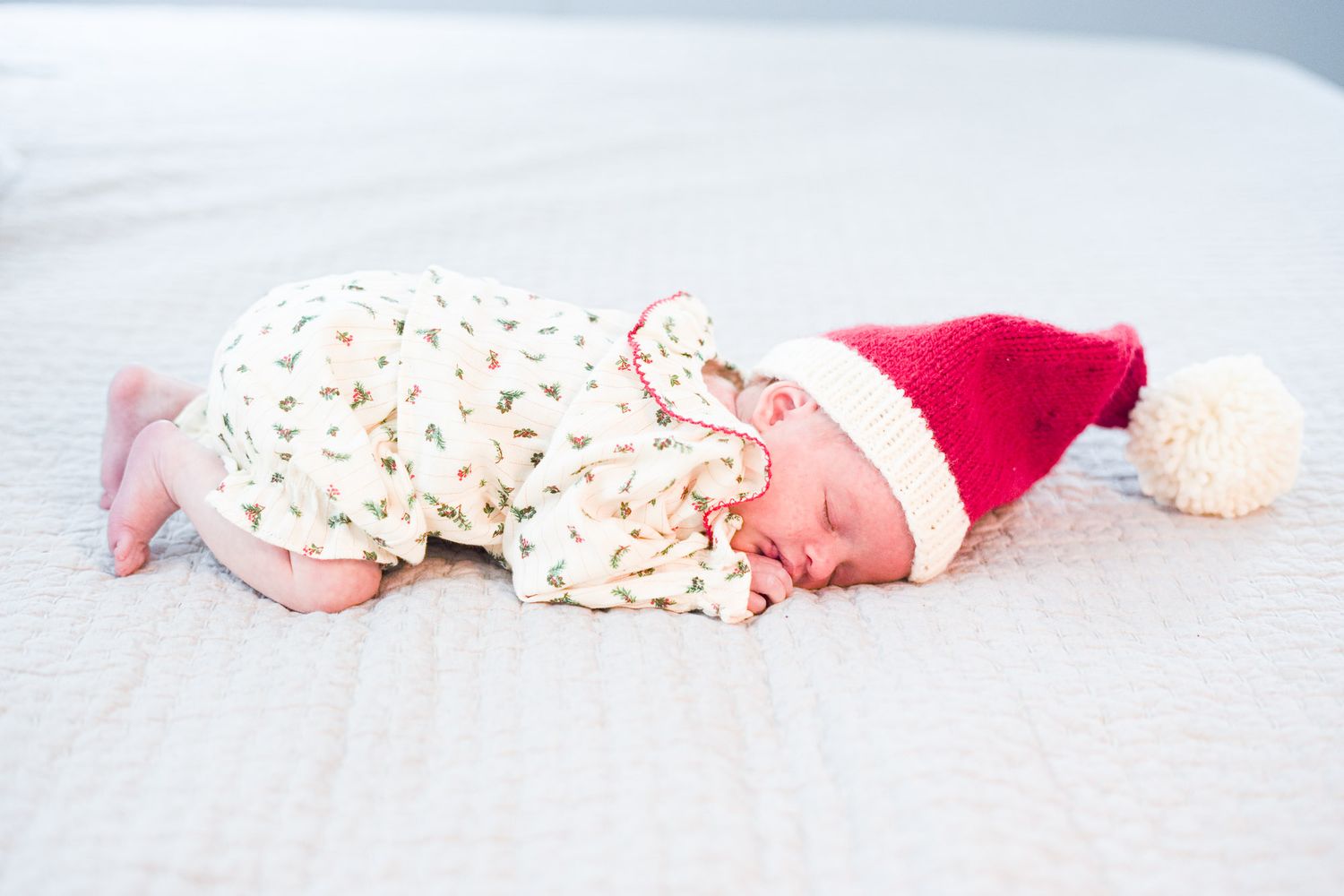 Sleeping newborn in Christmas tree print onesie wearing red Santa hat with cream pom-pom.