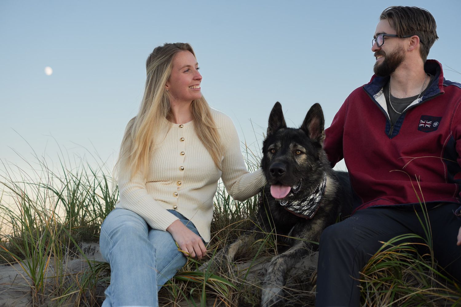 A couple sitting in beach grass with their German Shepherd dog under a clear sky.