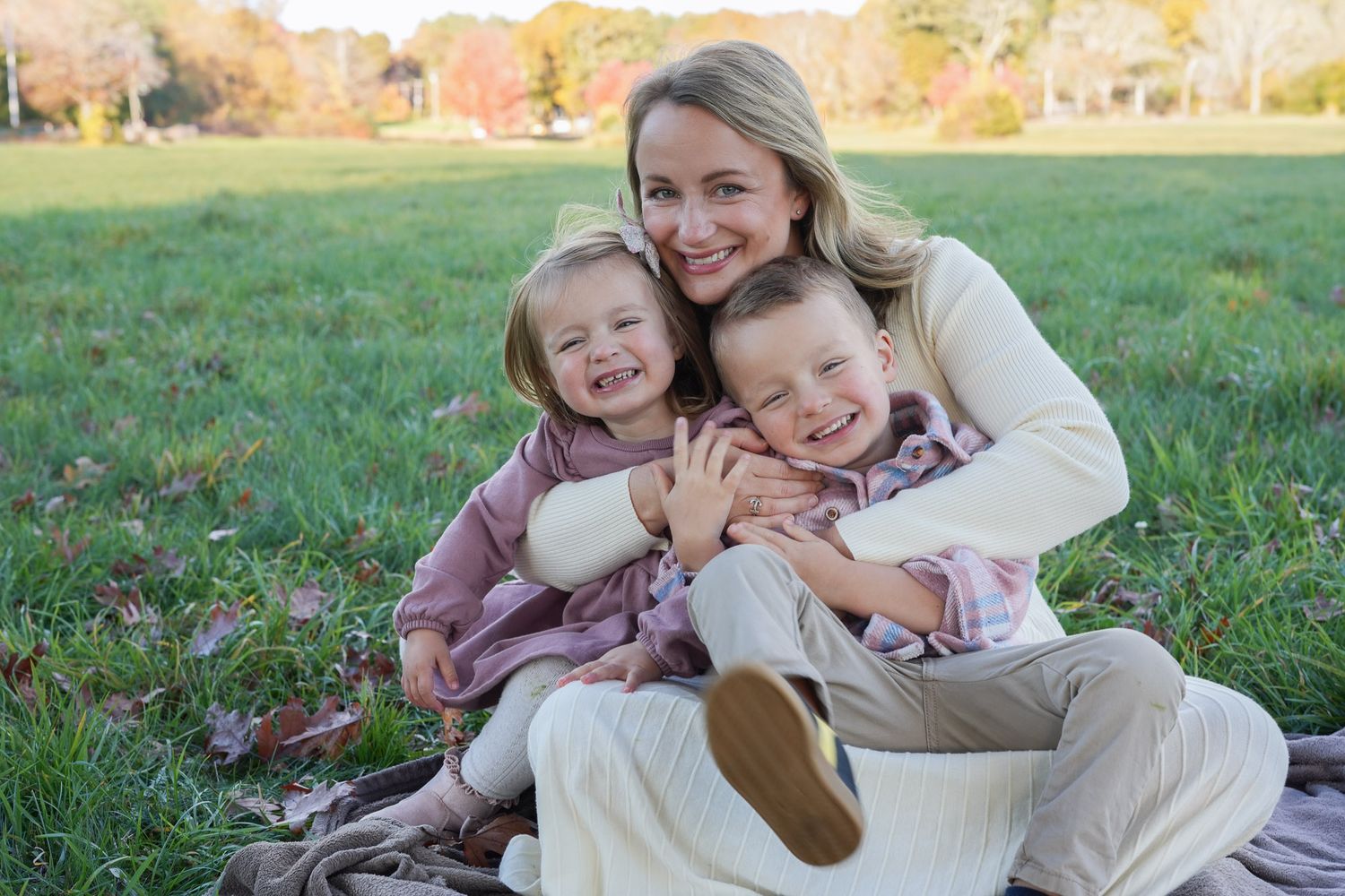 Smiling mother sitting on grass with two young children during an outdoor family photo session.