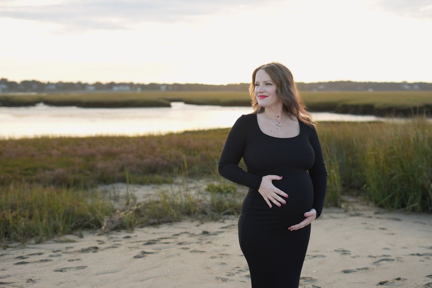 Pregnant woman in black dress cradling belly on sandy beach with marsh at dusk.