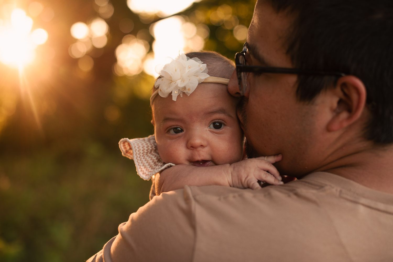 A father tenderly kisses his infant baby with a floral headband in warm golden sunset light.