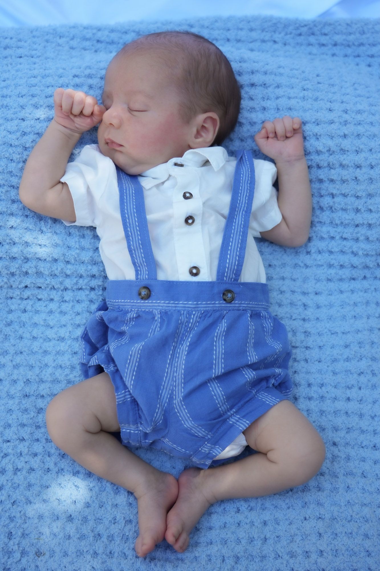 Peaceful sleeping newborn baby in blue suspender romper on blue knit blanket.