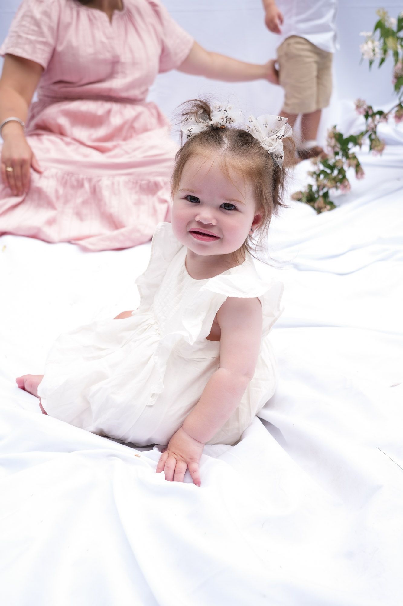 A cute baby girl in a white dress with a floral headband crawls on white fabric during an outdoor family photo session.