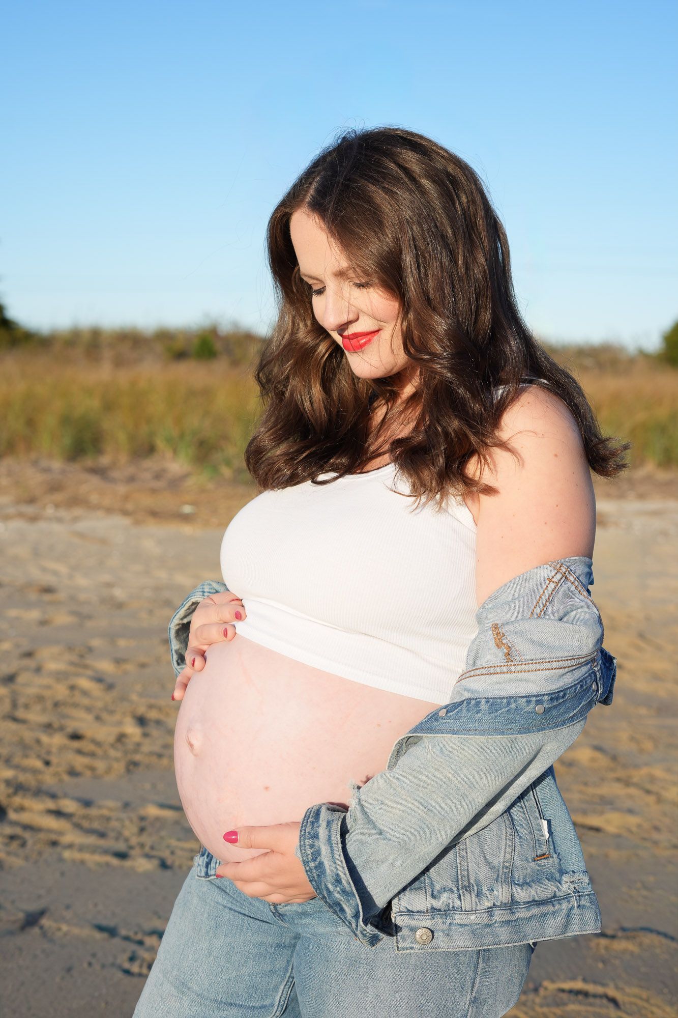 A pregnant woman with dark hair and red lips cradles her bare baby bump on a sunny beach.