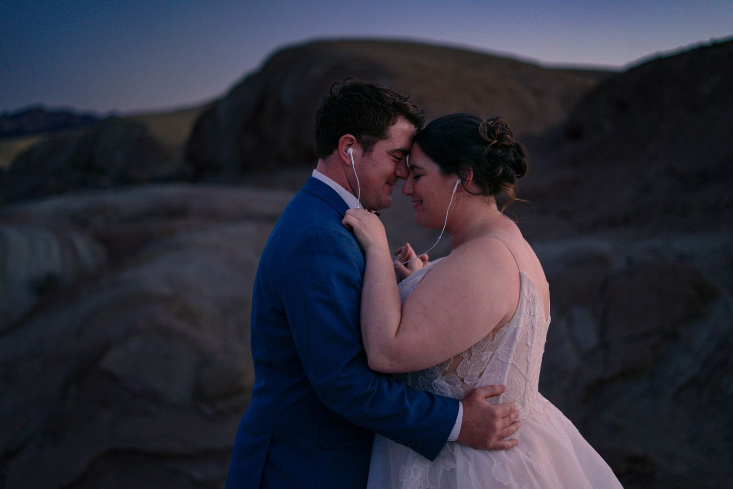Bride and groom sharing intimate moment forehead to forehead at twilight on rocky desert terrain.
