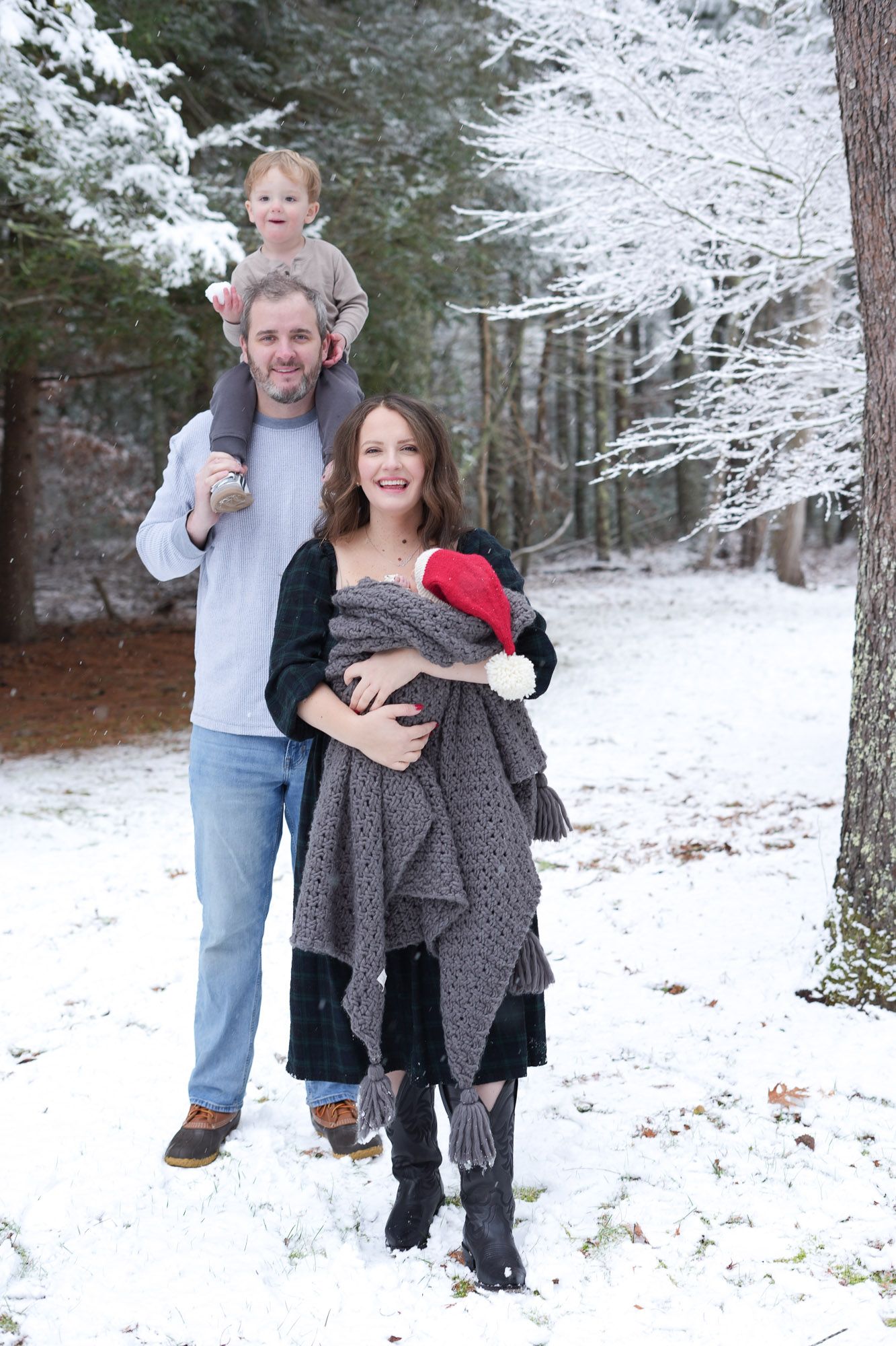 A smiling family of four poses together in a snow-covered forest, dad carrying toddler on shoulders while mom holds the newborn.