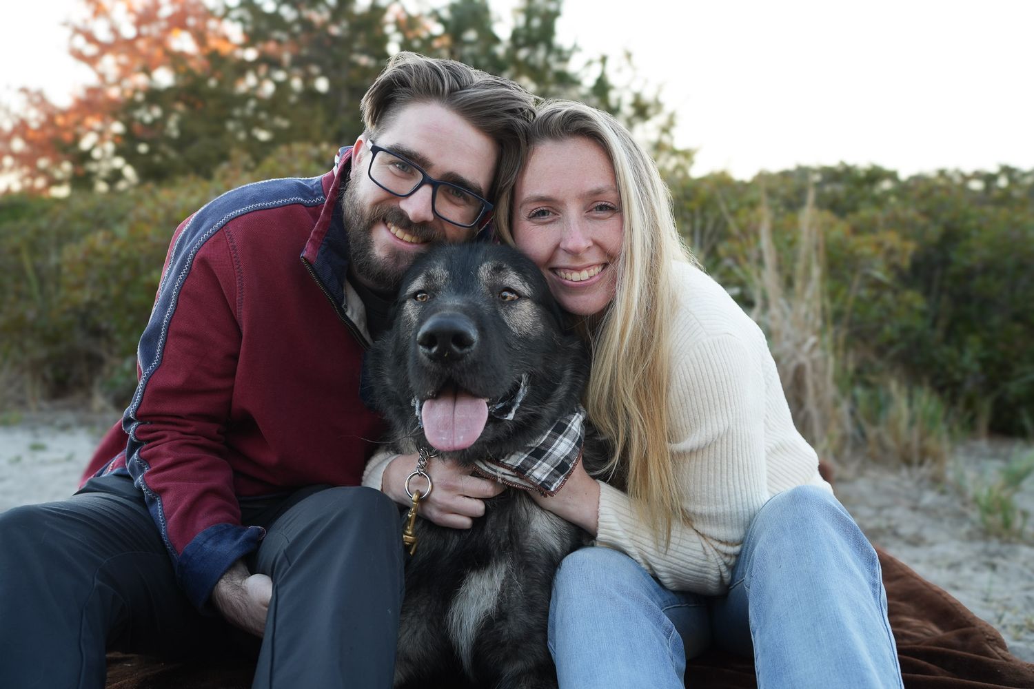 Happy couple hugging their German Shepherd dog while sitting on a beach at golden hour.