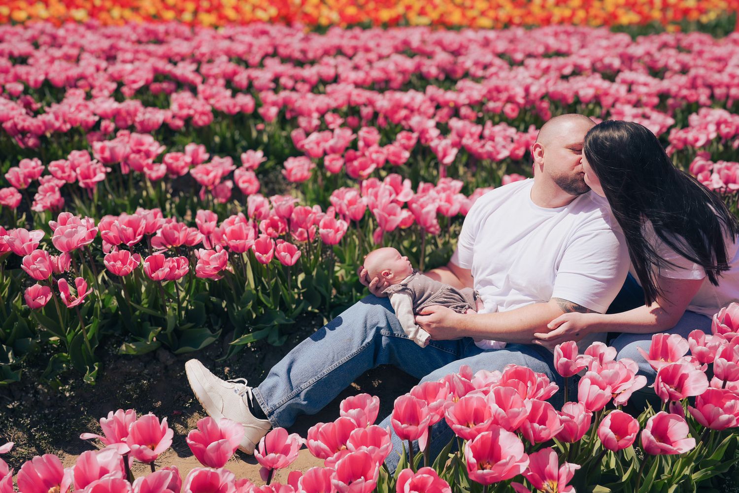 A couple sits closely together in a vibrant pink tulip field, cradling their newborn baby.