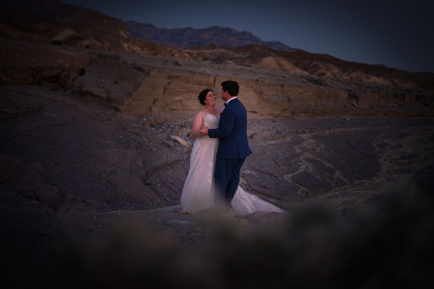 Bride and groom dancing together at twilight among dramatic desert rock formations and mountains.