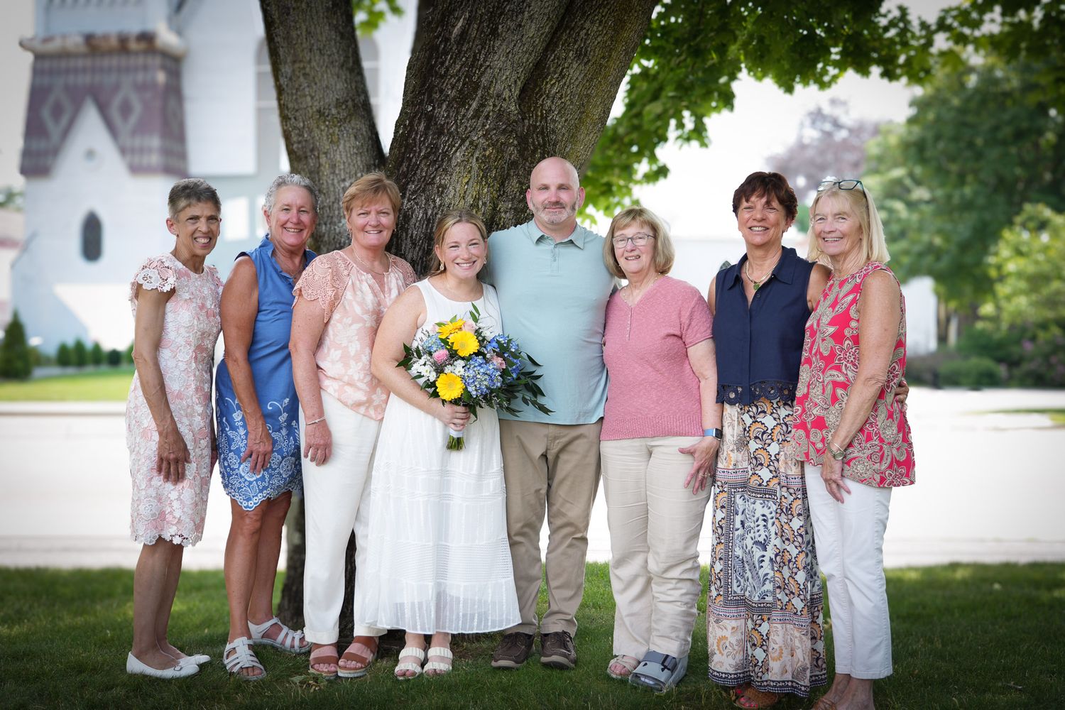 A group of seven adults smiling together outdoors near a large tree with a white church in background.