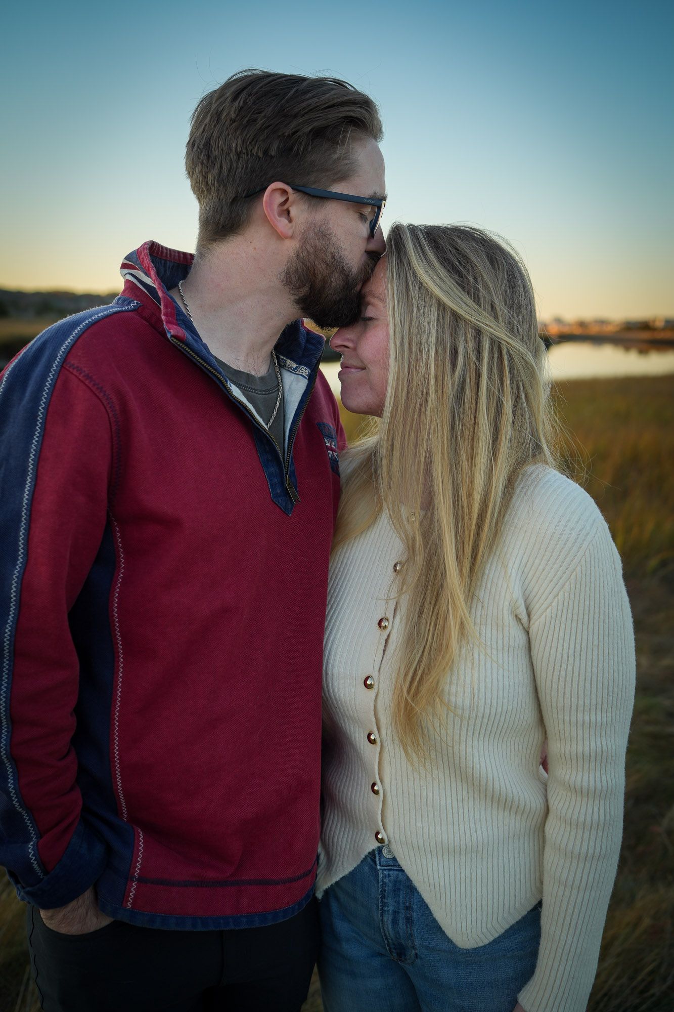 Couple sharing a tender forehead kiss at sunset in a marsh field, man in red jacket and woman in cream sweater.