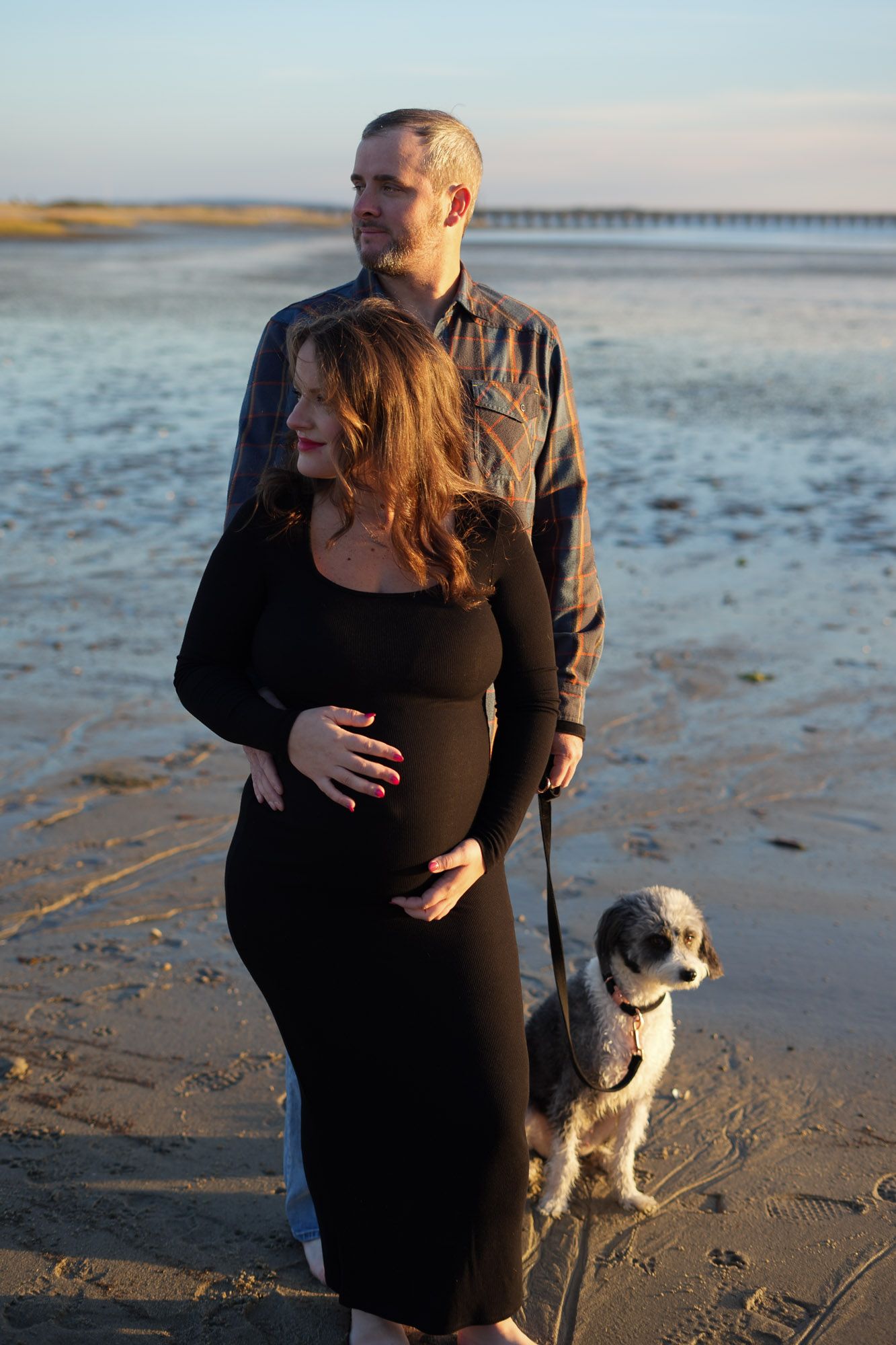 Expectant couple with dog on leash standing on beach at sunset, woman in black dress.
