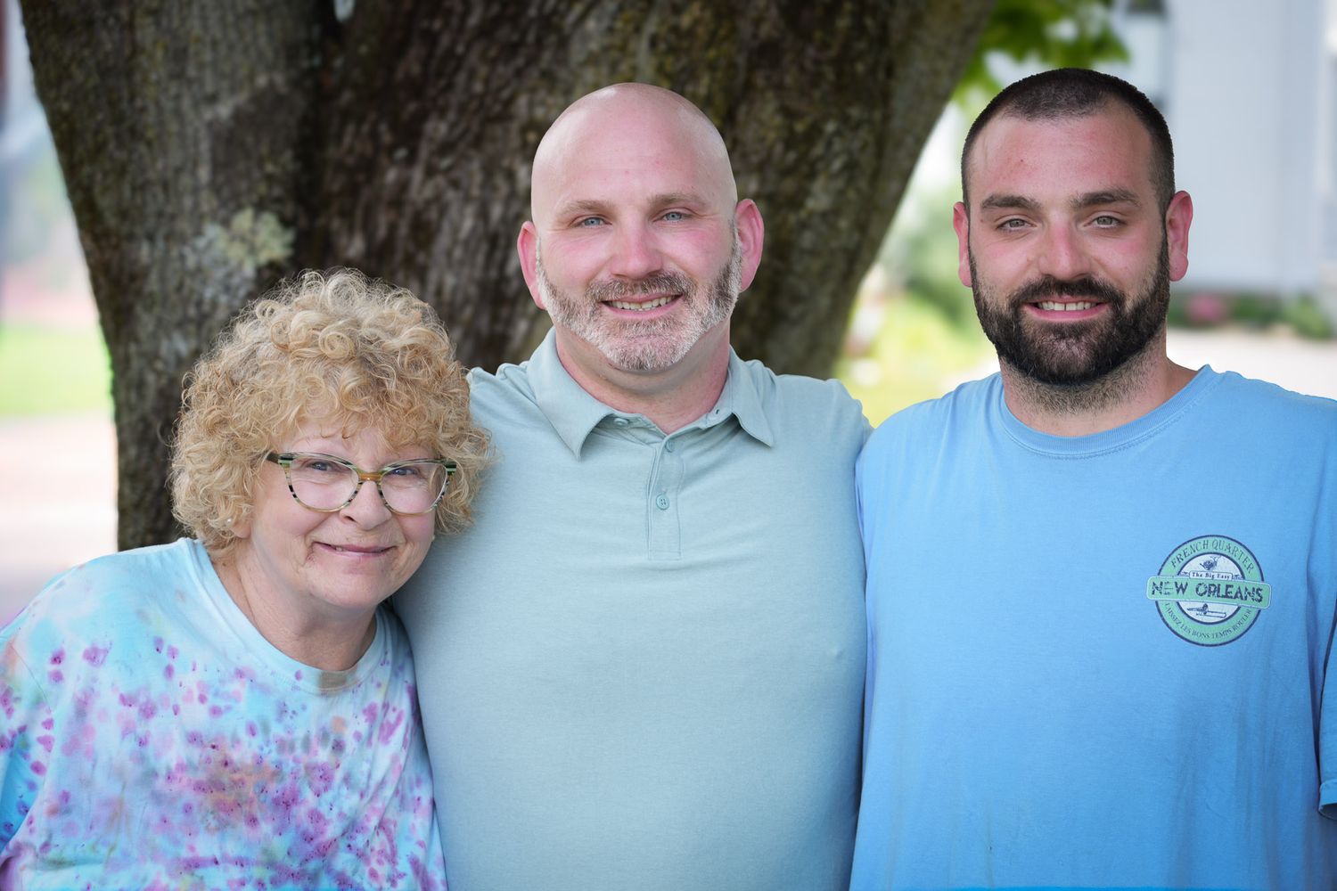 Three people posing together outdoors under a tree, smiling for a family photo at a wedding celebration.