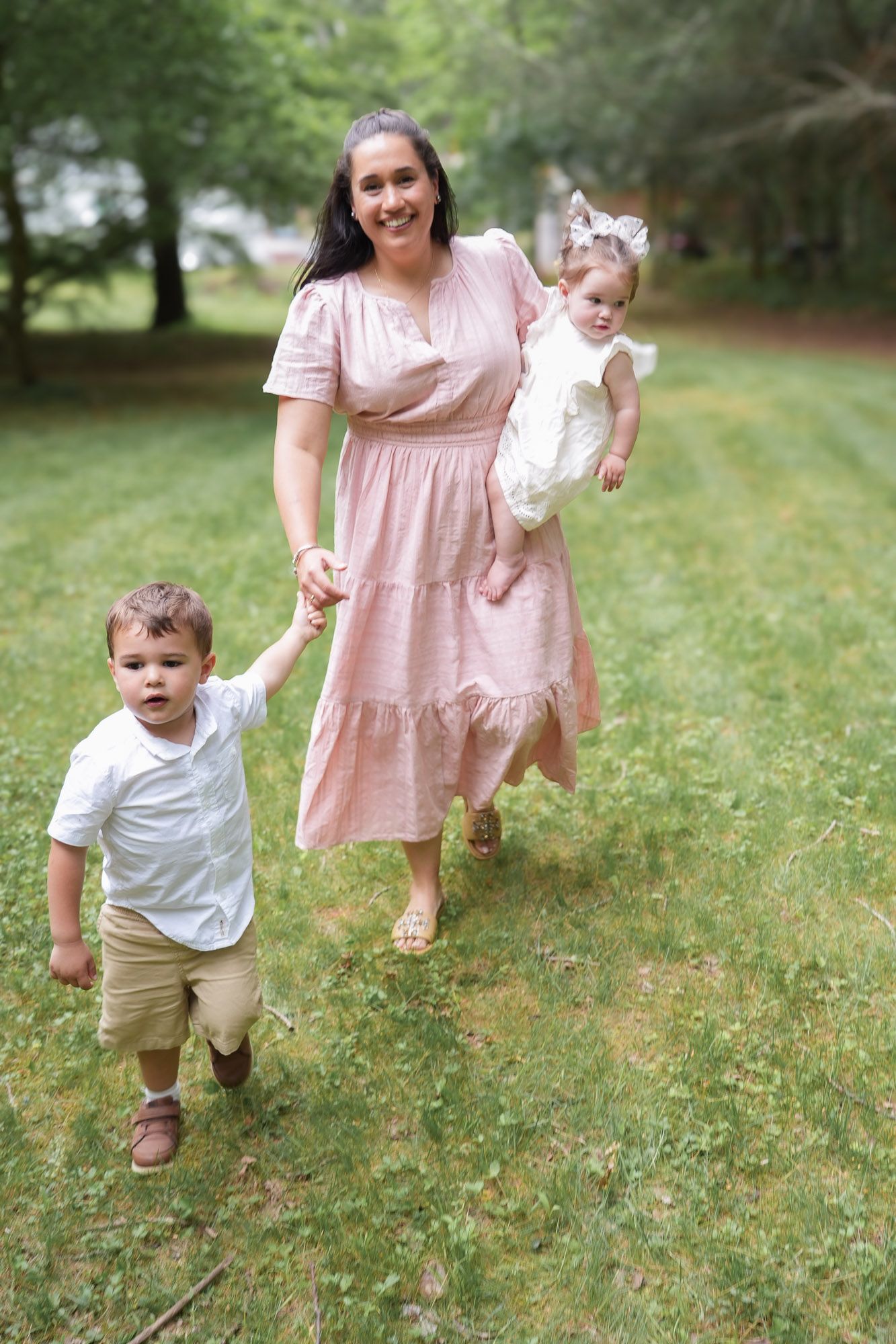 Mother in pink dress walking with young son and baby daughter in a green outdoor setting.