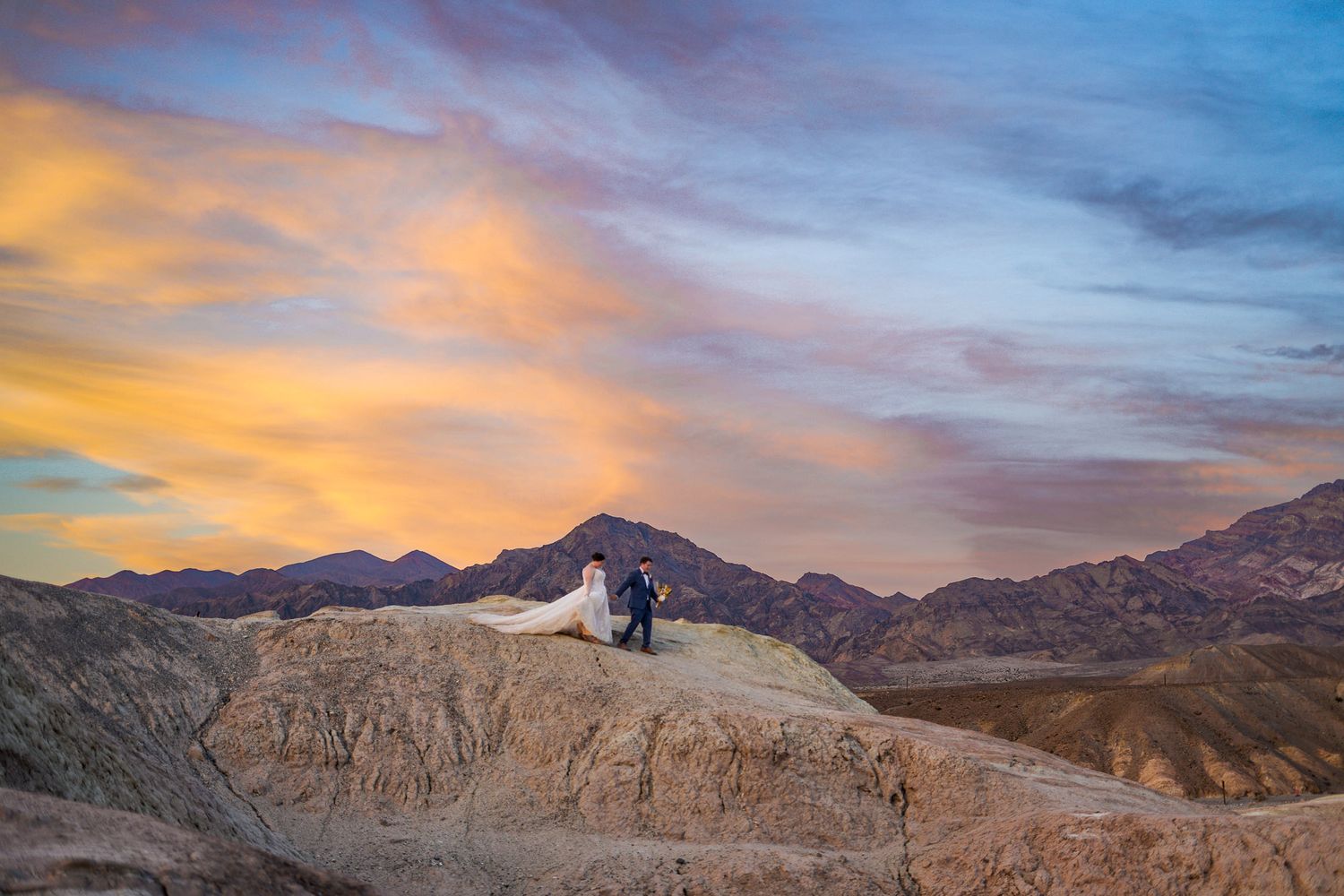 Wedding couple silhouetted against a vibrant orange and blue sunset sky atop rocky Death Valley desert formation.