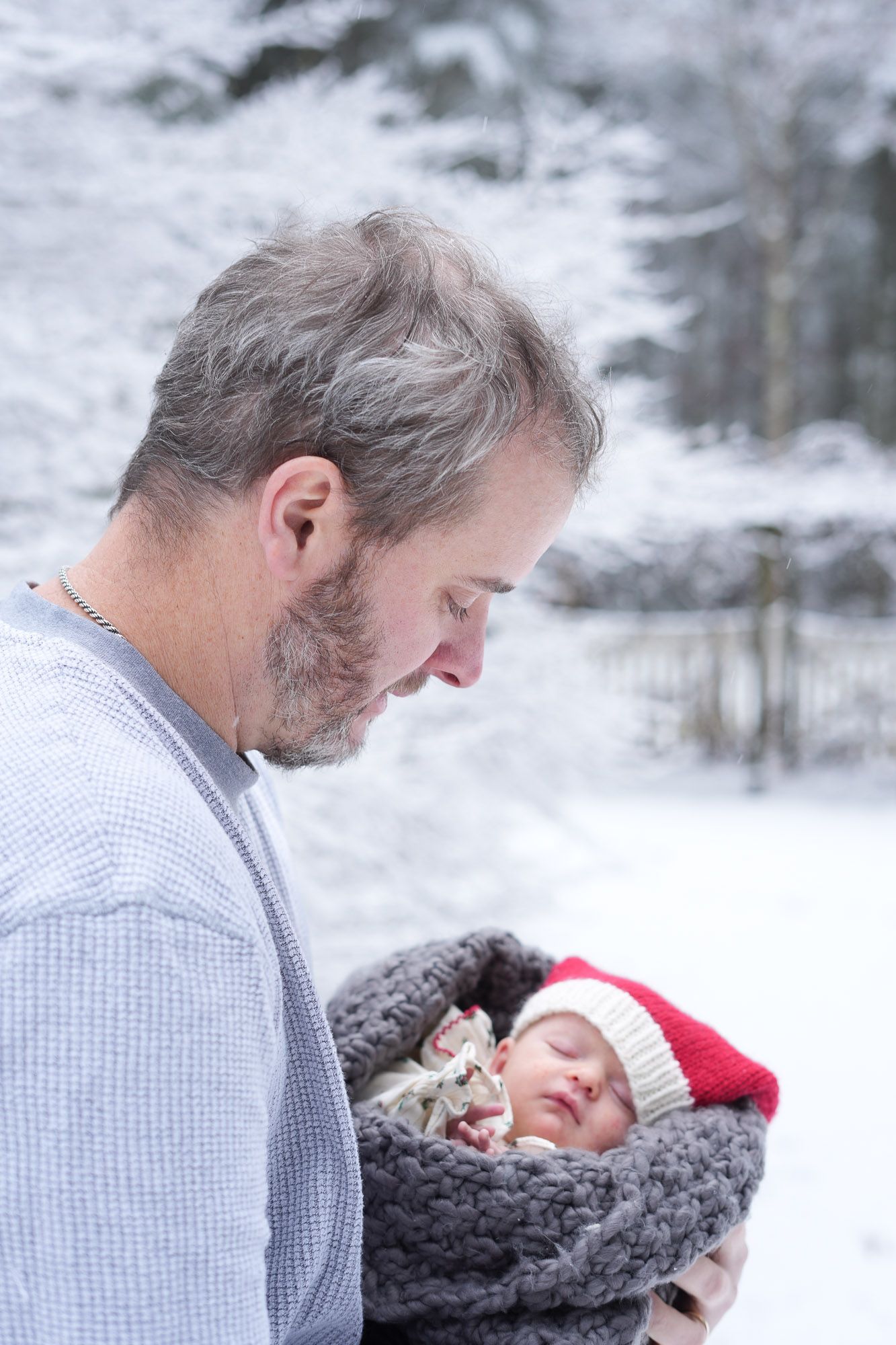 Loving father gazing down at sleeping newborn baby in red Santa hat wrapped in gray knit blanket in snow.