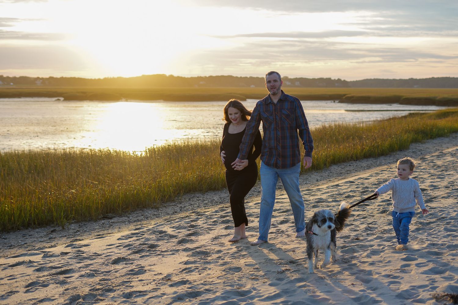 Family walking on sandy beach at sunset with dog on leash, pregnant mother and father with young child.