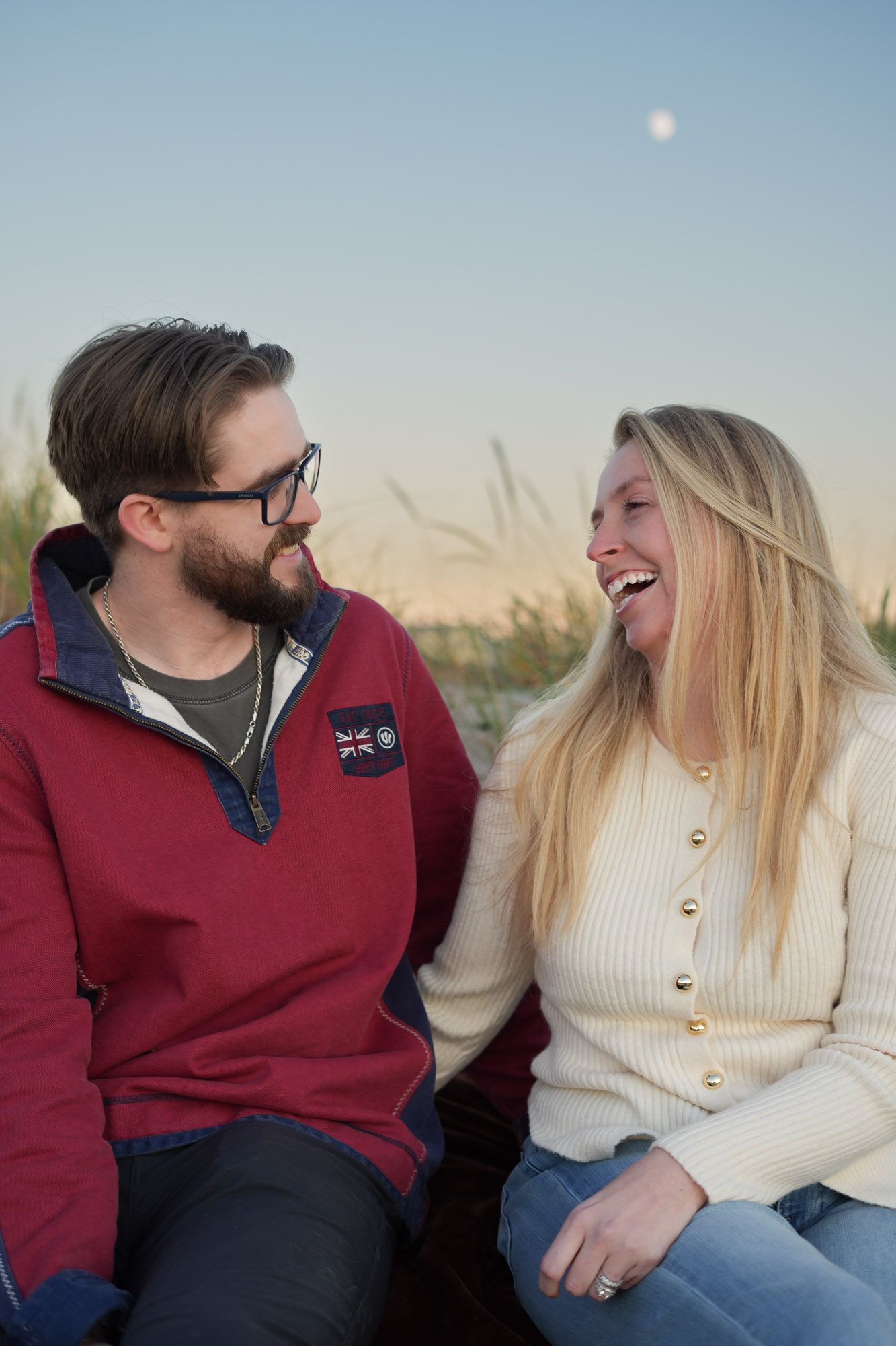 A laughing couple sitting together outdoors at dusk with the moon in the background.