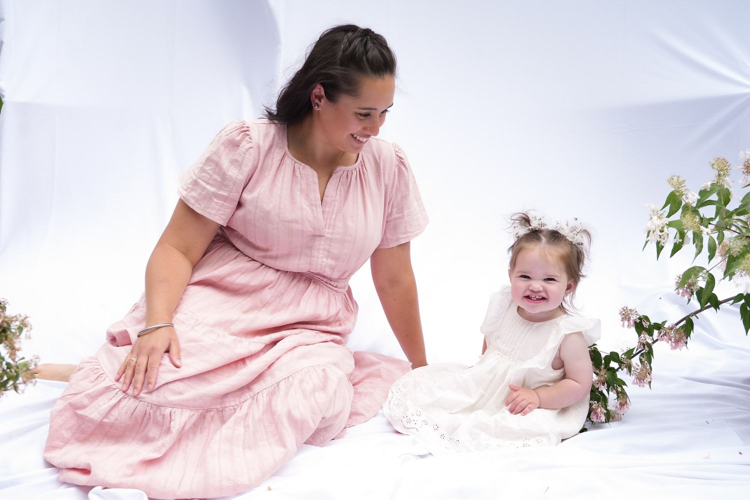 A smiling mother in a pink dress plays with her happy toddler daughter in a white dress on a white backdrop.