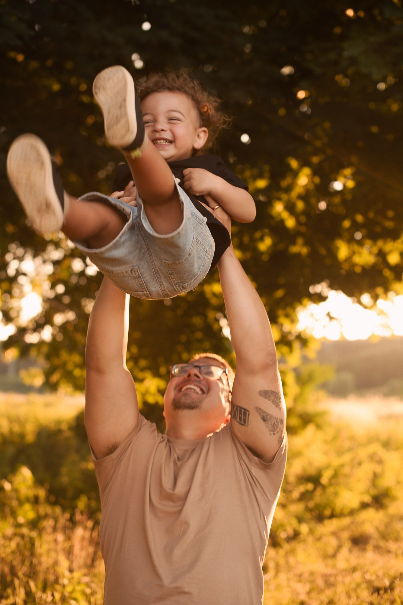 A joyful father lifts his laughing toddler high in the air during a golden sunset outdoor photo session.