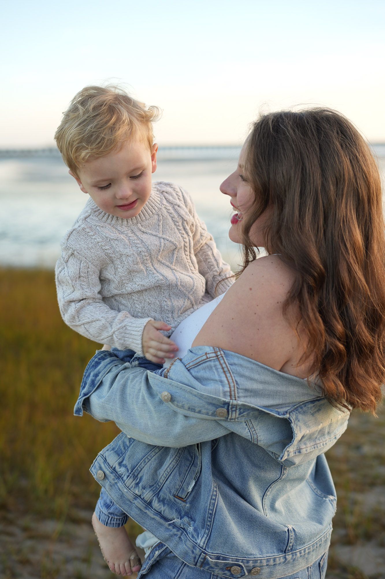 Pregnant woman in denim jacket holding smiling toddler on beach at golden hour.