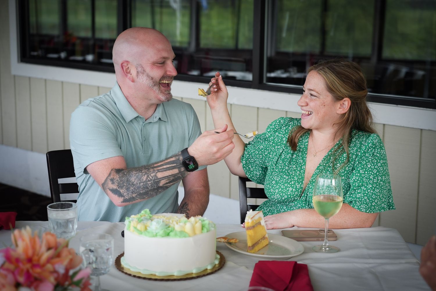 Happy couple sharing cake at wedding reception, man with tattoos feeding woman in green floral dress.