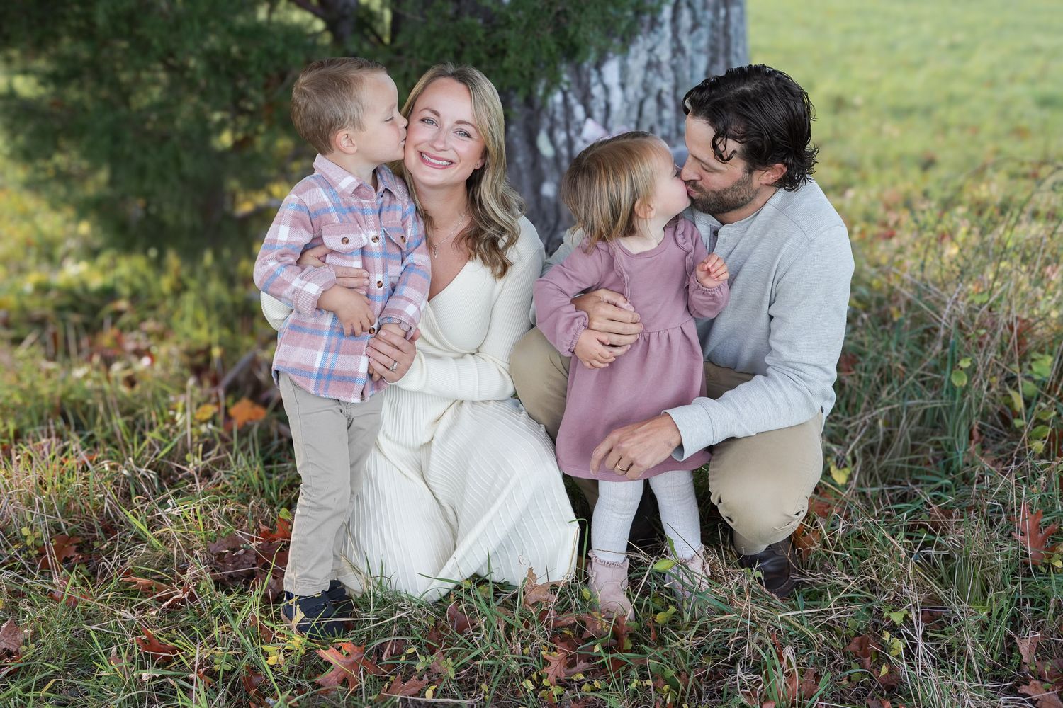 Family of four sitting under a tree as children kiss their parents on the cheeks outdoors.
