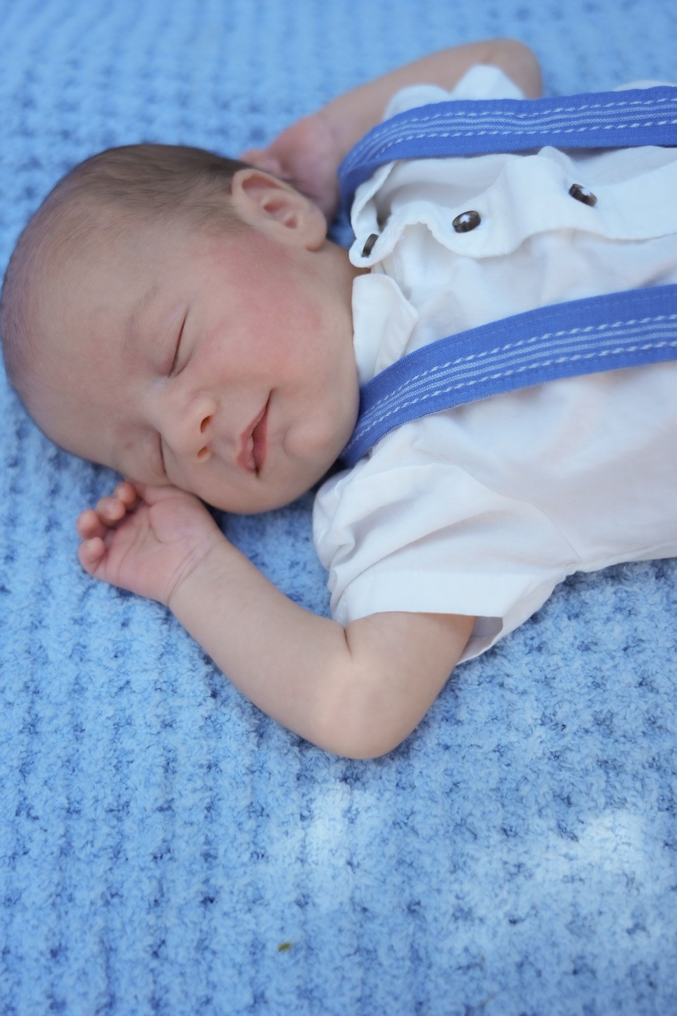 Peaceful sleeping newborn baby in white shirt with blue suspenders on knit blanket.
