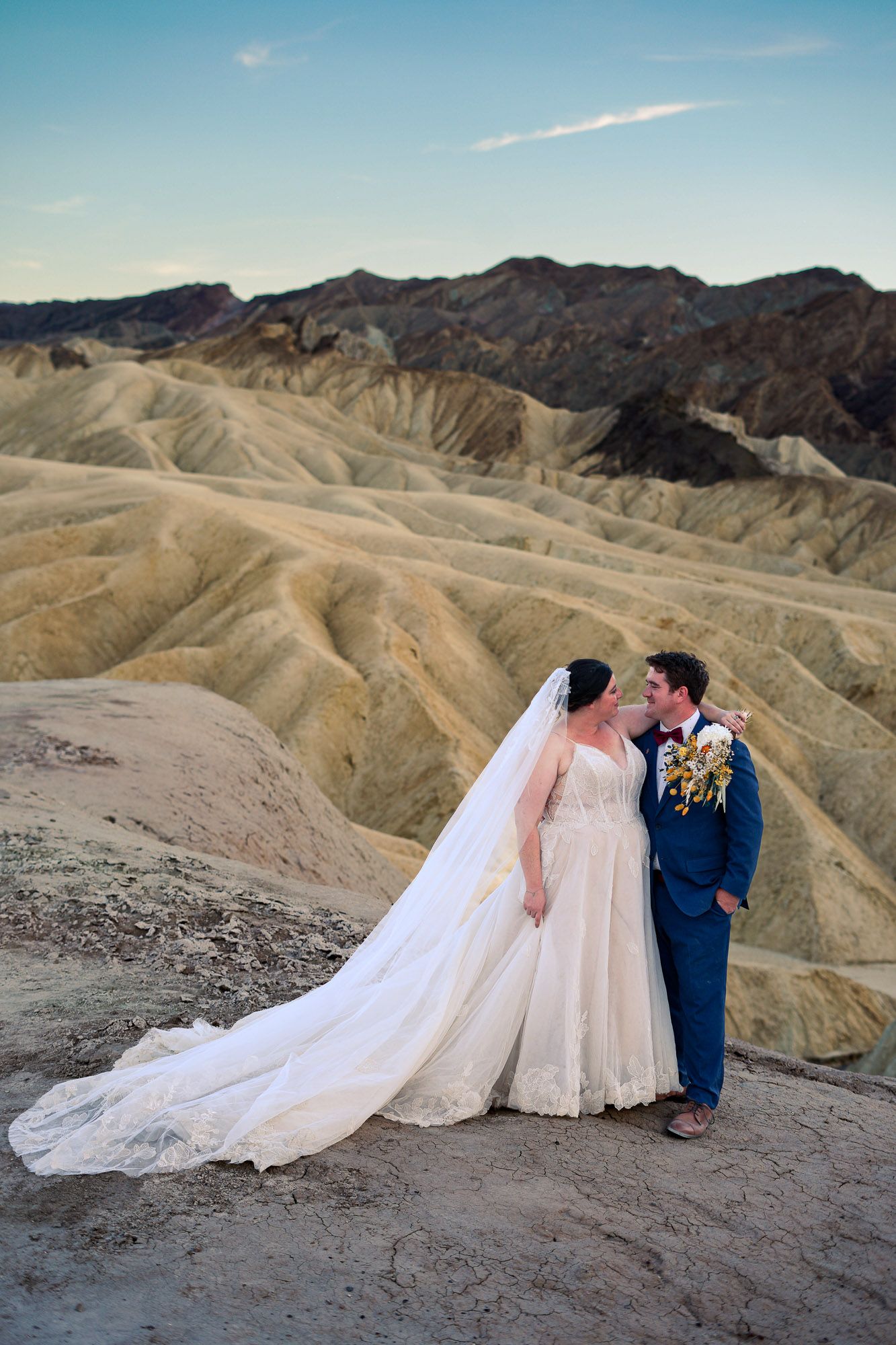 Bride in lace gown with long veil and groom in blue suit pose at golden Zabriskie Point badlands.