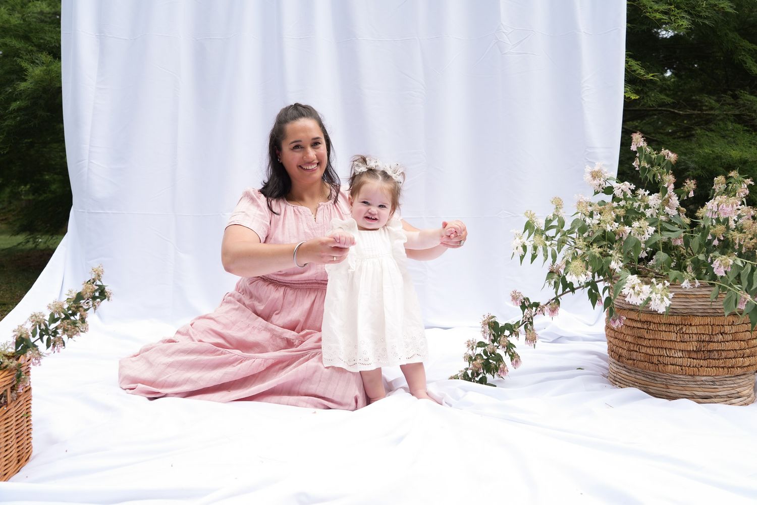 A mother in a pink dress sits on white fabric smiling at her standing toddler in a white dress with floral decor.