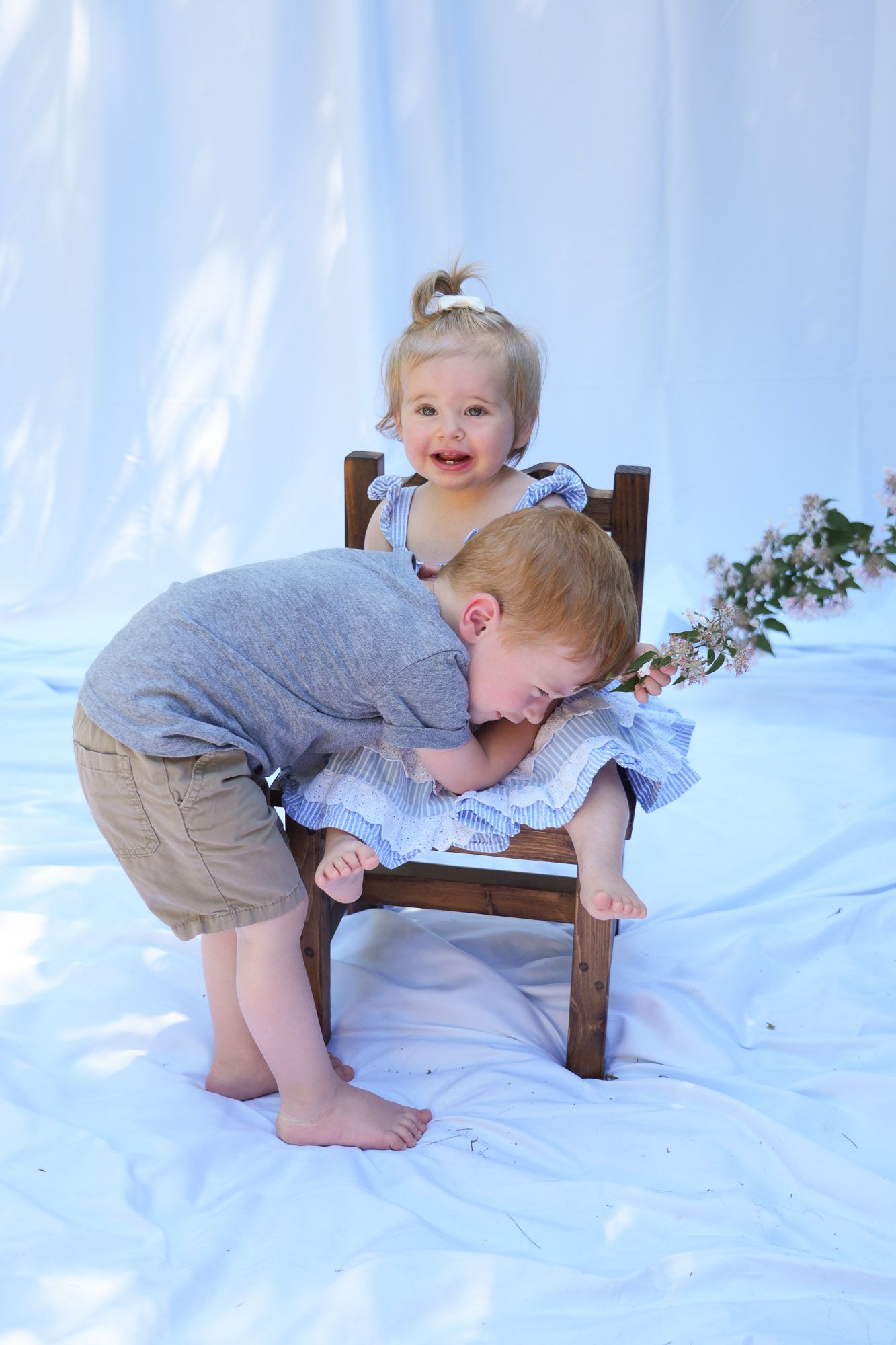 Two young children playing on wooden chair outdoors with white fabric backdrop.