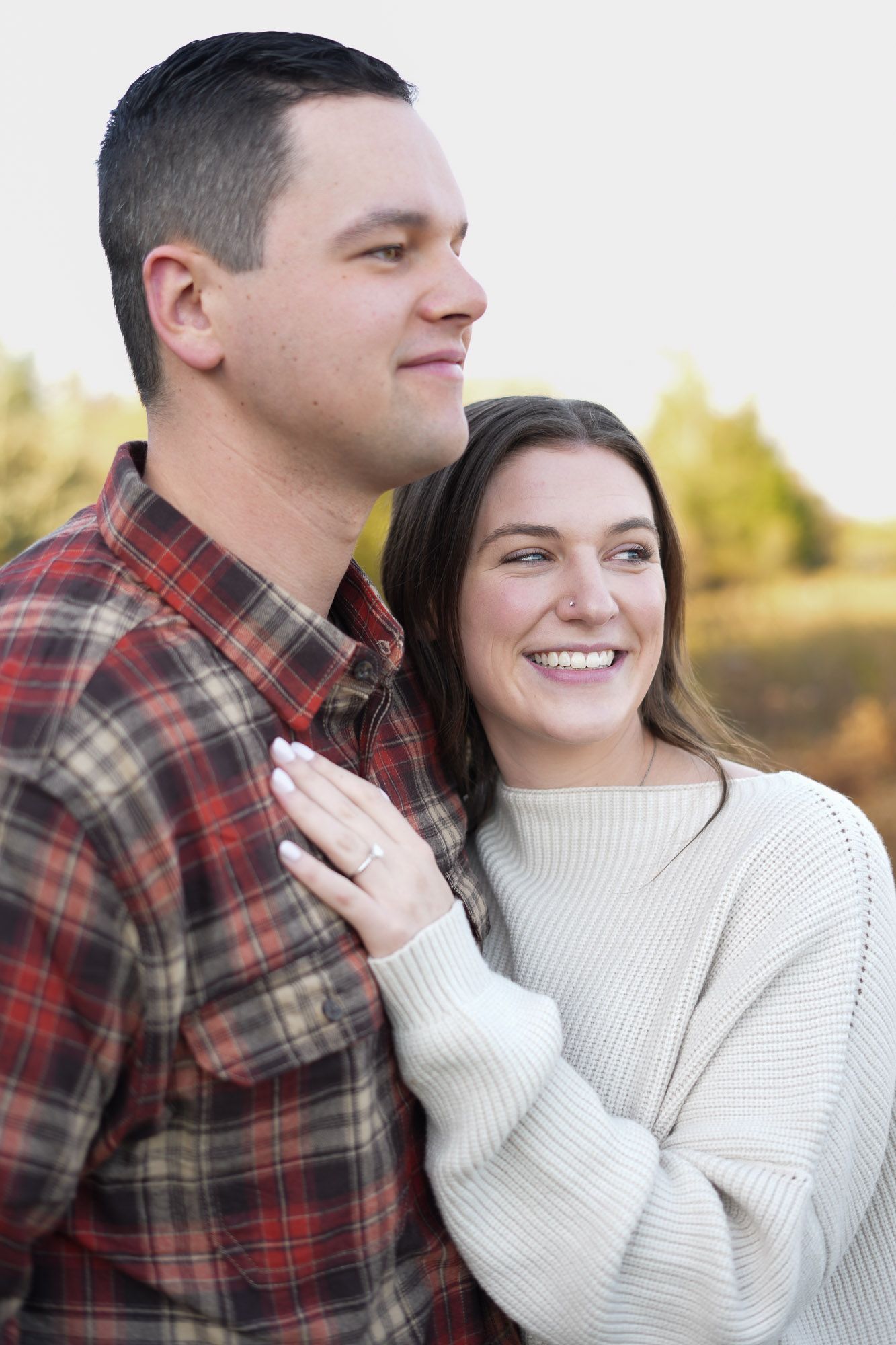 Smiling couple in autumn outdoor setting, woman in cream sweater embracing man in plaid flannel shirt.