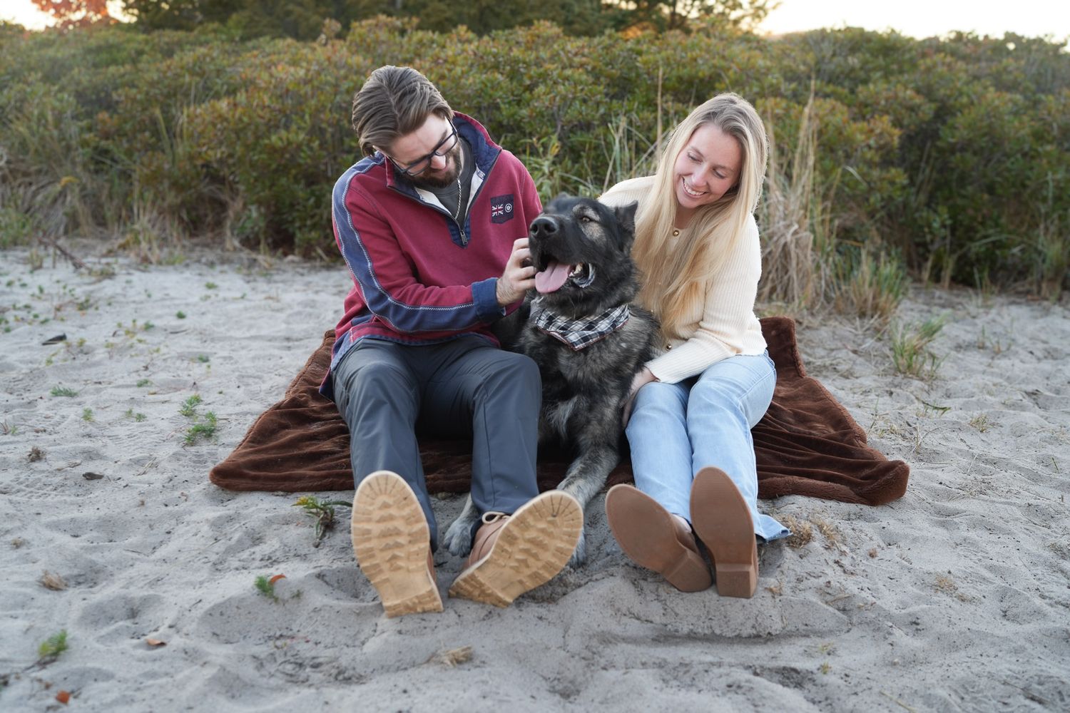 Couple sitting on brown blanket on sandy beach petting their large dark German Shepherd dog.