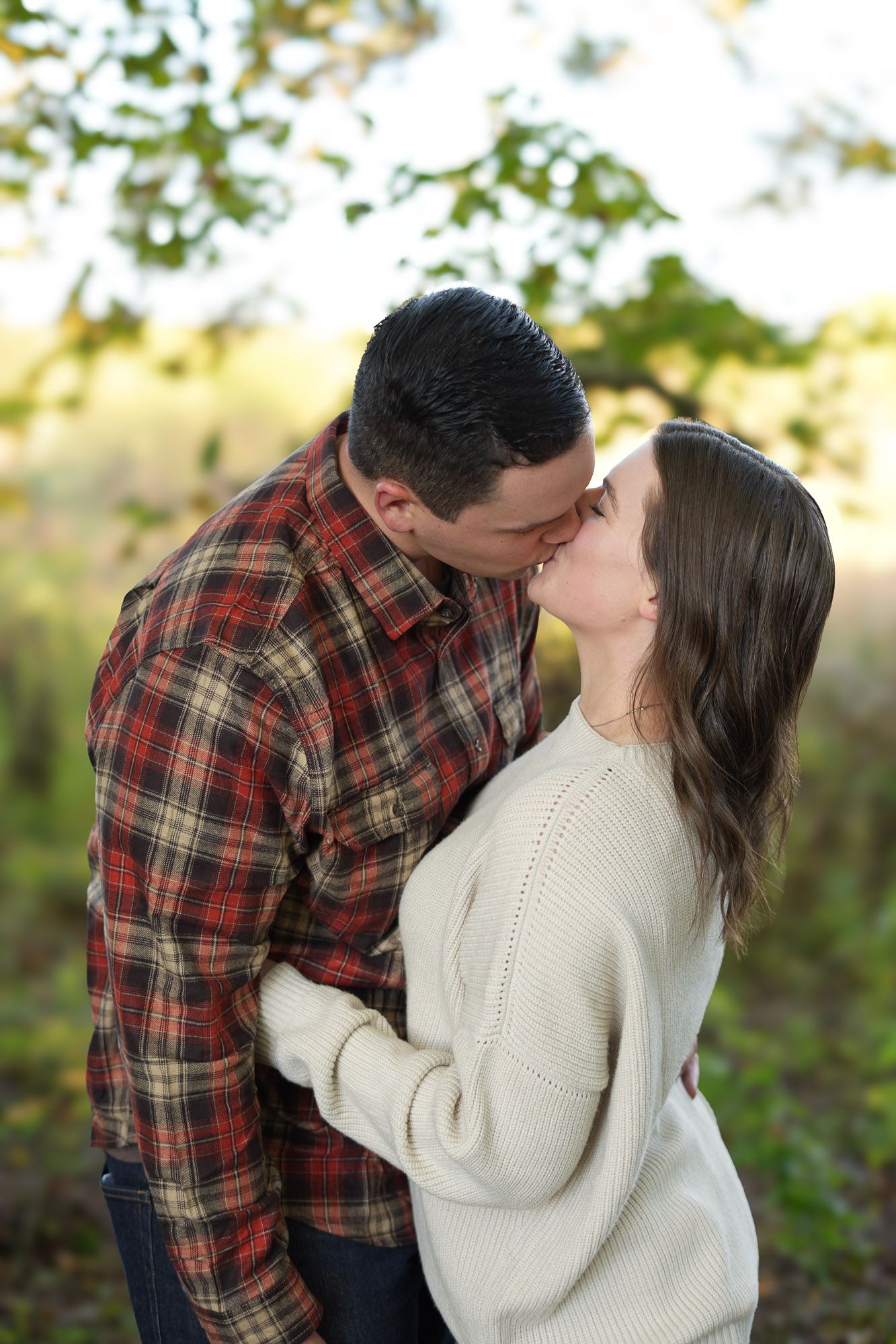 Couple sharing a tender kiss outdoors with soft bokeh autumn foliage in the background.