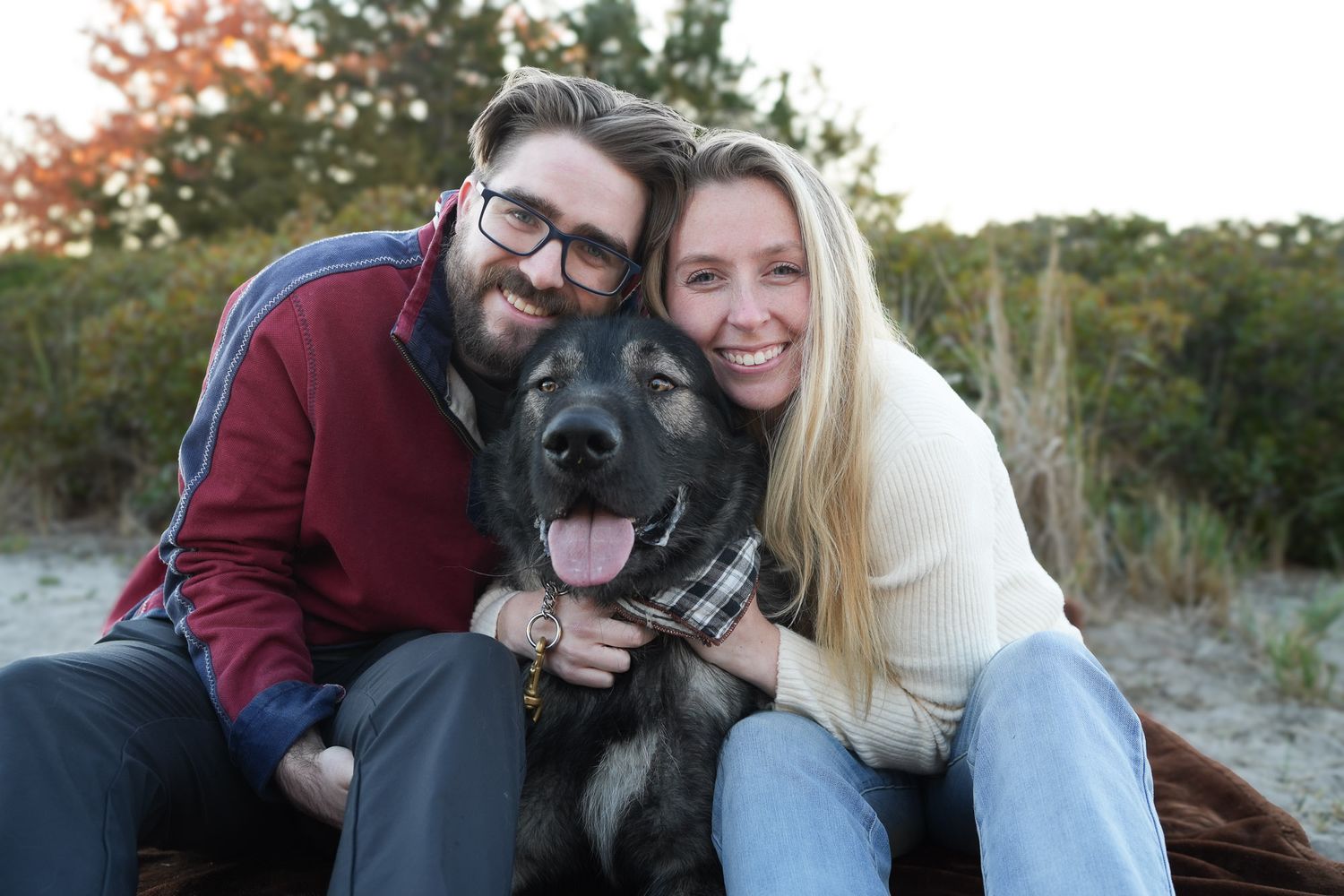 Happy couple hugging their German Shepherd dog with plaid bandana on a sandy beach.