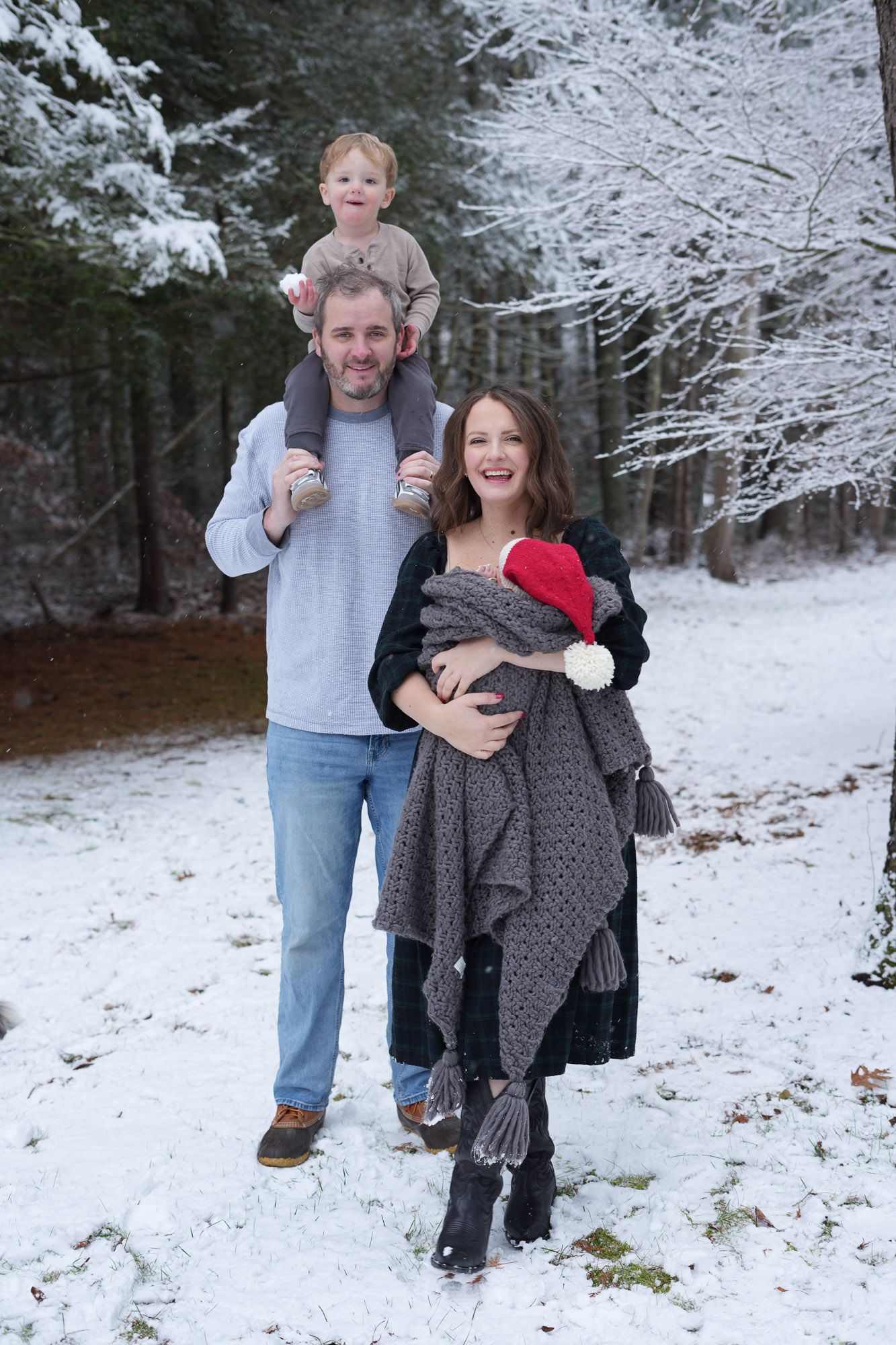 Happy family posing in snowy forest, dad carrying toddler on shoulders, mom holding newborn.
