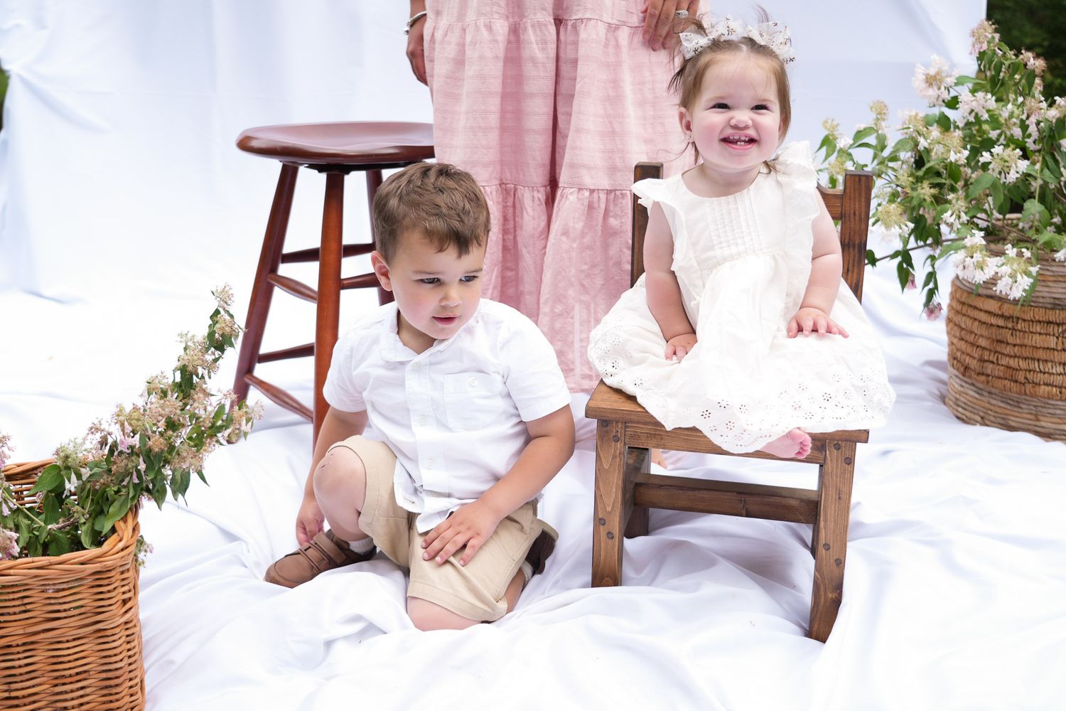 Two young children in white outfits sit together outdoors on white blanket with wicker flower baskets behind them.