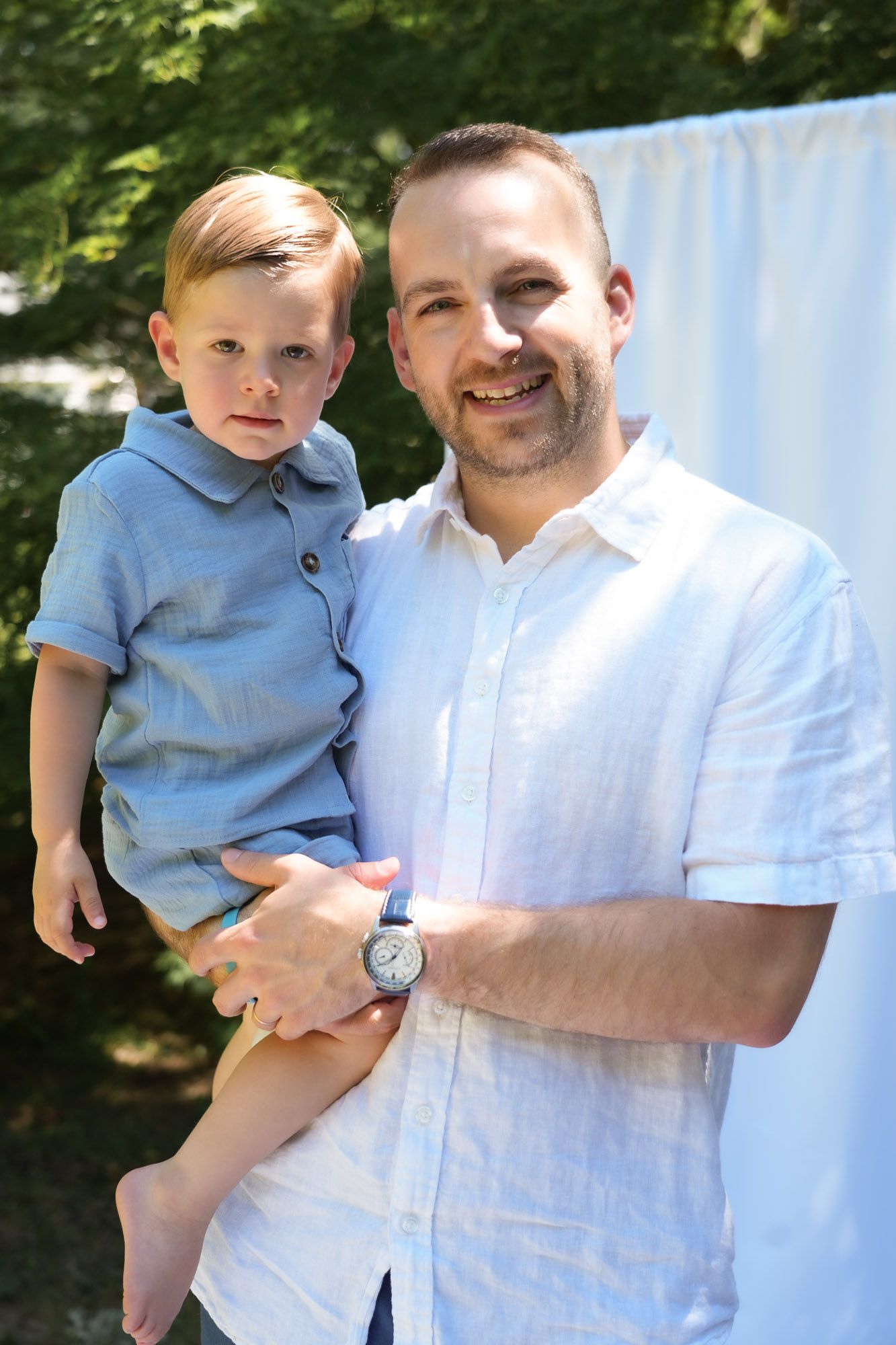 Smiling father holding his young blonde son in a blue shirt in an outdoor portrait.