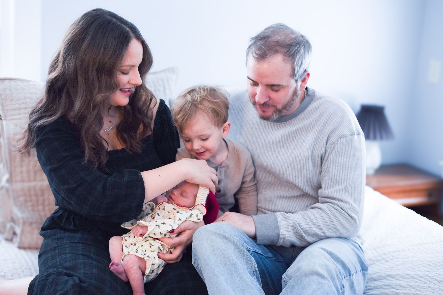 Happy family of four sitting on bed with parents and toddler admiring their newborn baby.