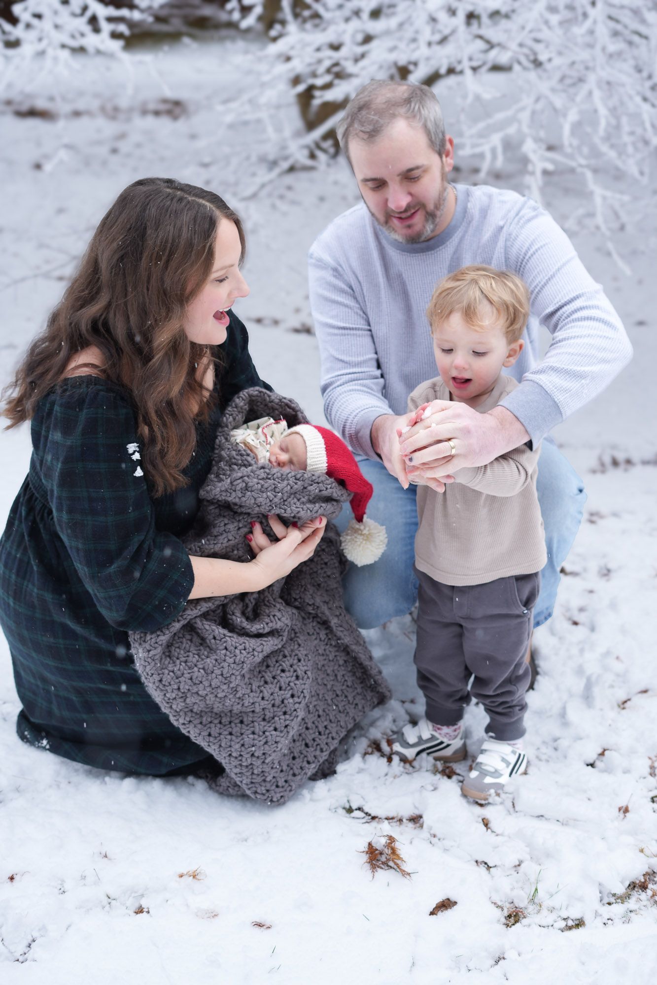 A happy family of four kneels in the snow, parents smiling as their toddler son meets their newborn baby for the first time.
