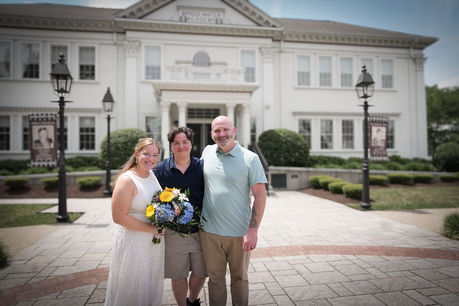 Bride, officiant, and groom posing together in front of large white building after ceremony.