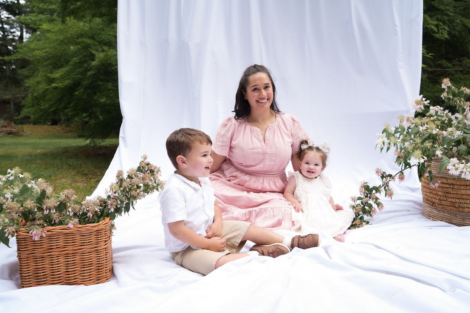 A smiling mother in a pink dress poses with her two young children on white fabric with flower baskets in an outdoor setting.