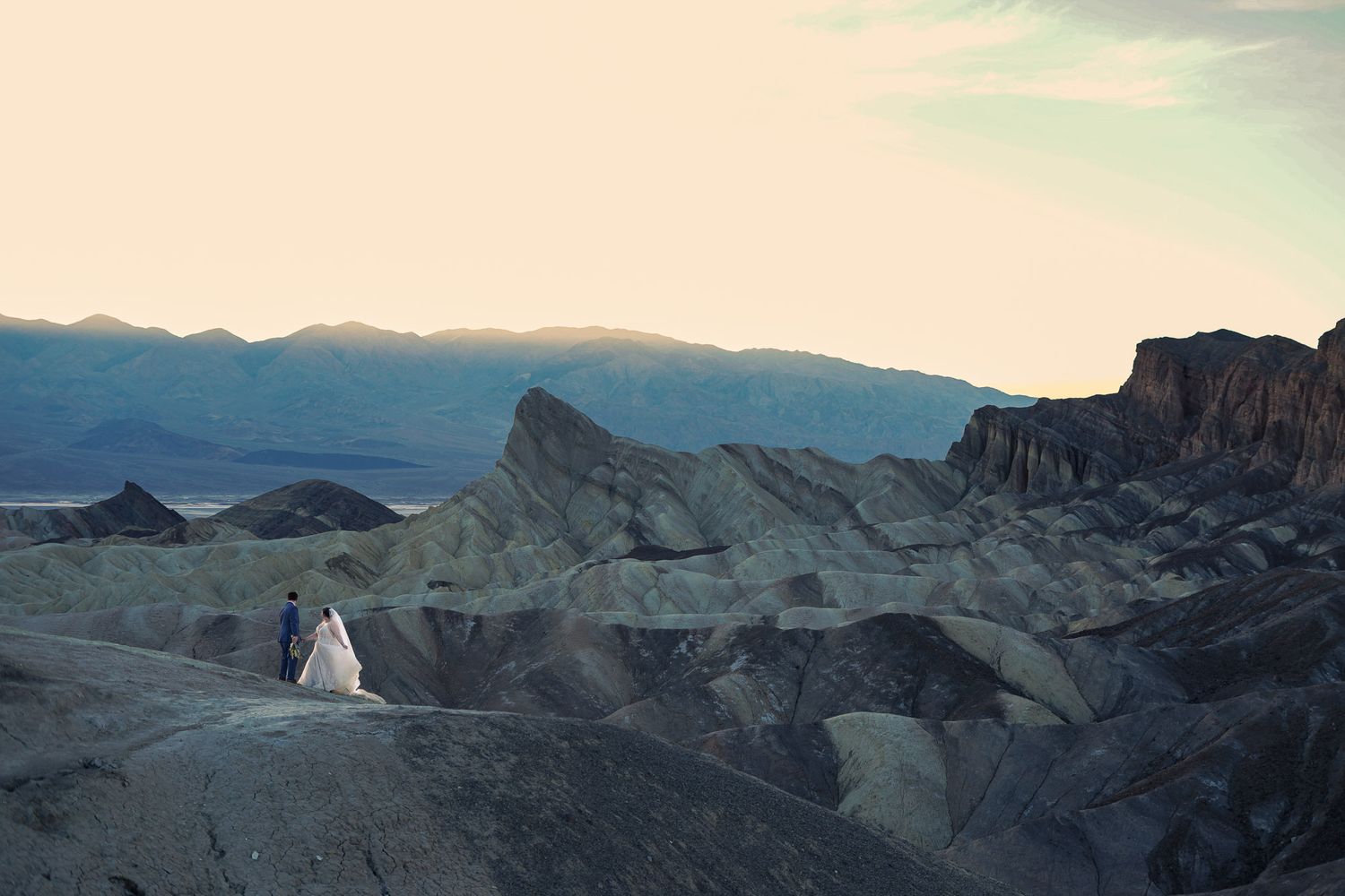 Wedding couple walks along rugged Death Valley ridge at dusk with mountain silhouettes in background.