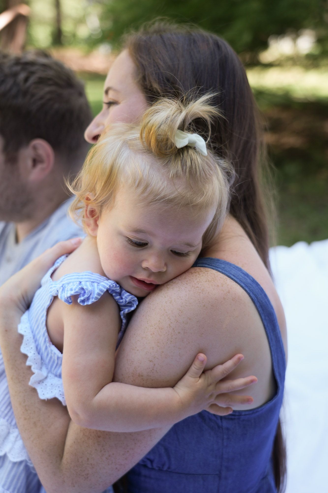 A mother holds her sleepy blonde toddler girl in a blue ruffled dress outdoors.