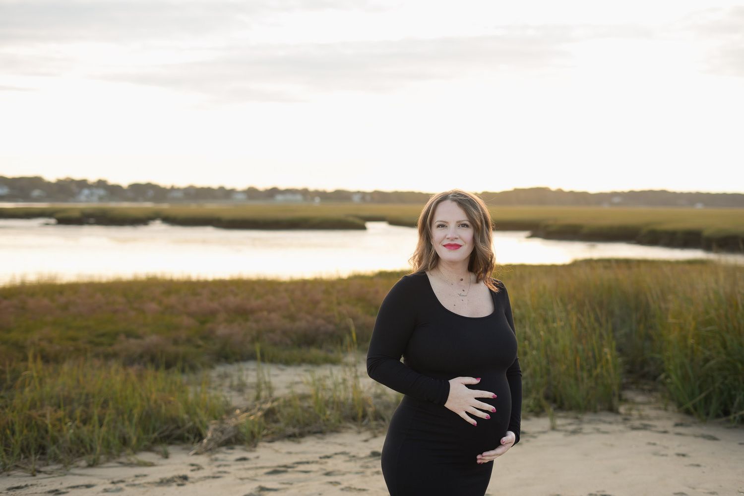 Pregnant woman in black outfit smiling on sandy beach with golden marsh at sunset.