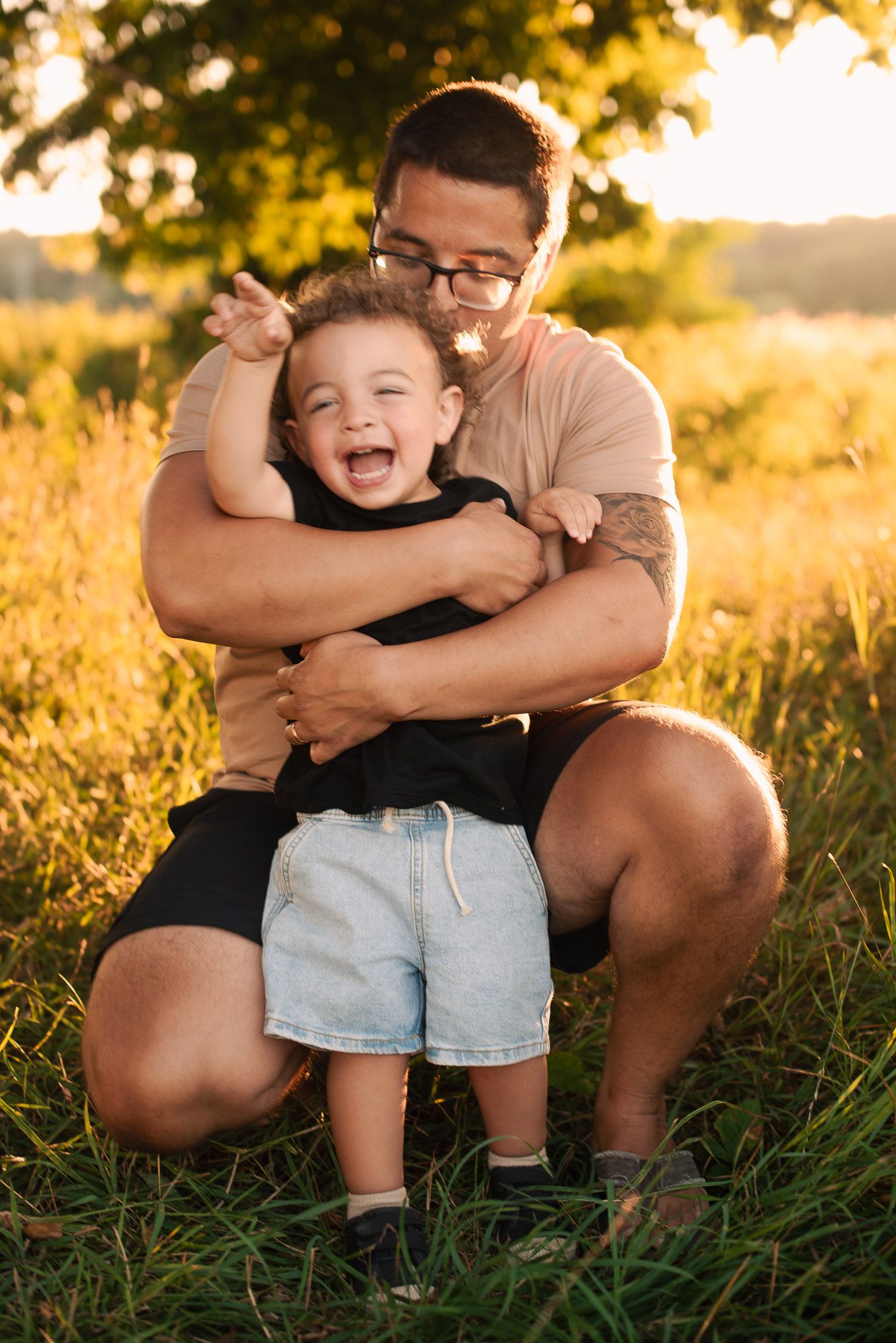A father hugs his giggling young son in a grassy field bathed in warm golden sunlight.