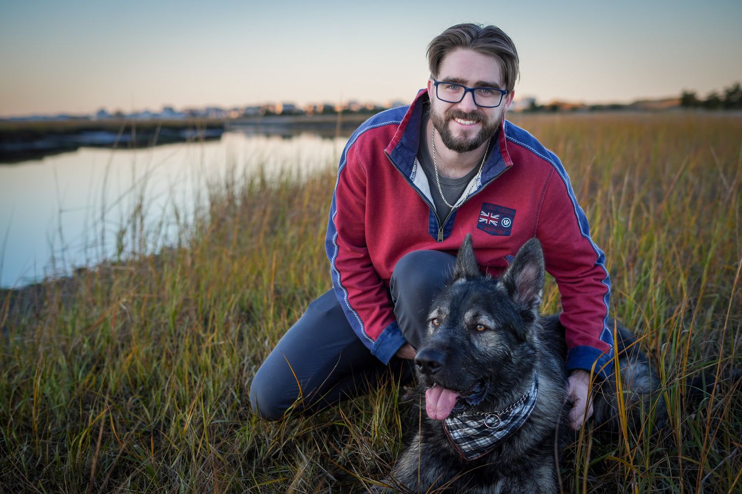 A smiling bearded man in a red jacket kneeling beside his German Shepherd near a marsh.