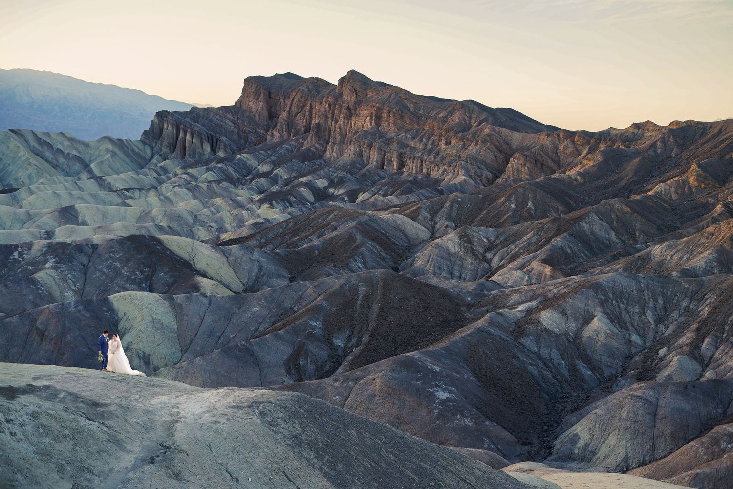 Bride in white gown and groom stand together in sweeping Death Valley desert mountain terrain.